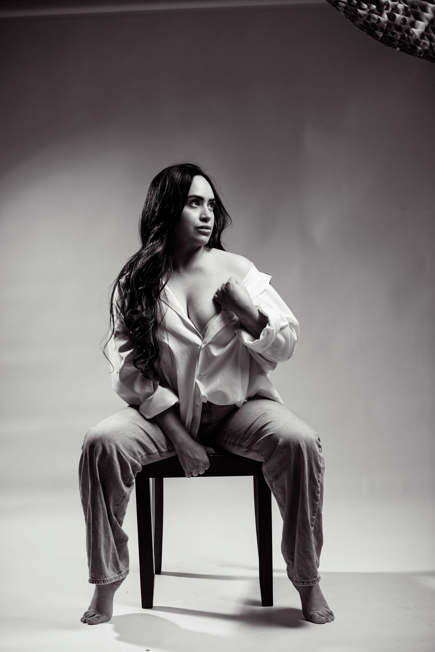 Black and white studio photo of a woman in an open white shirt and jeans sitting on a chair with long wavy hair.