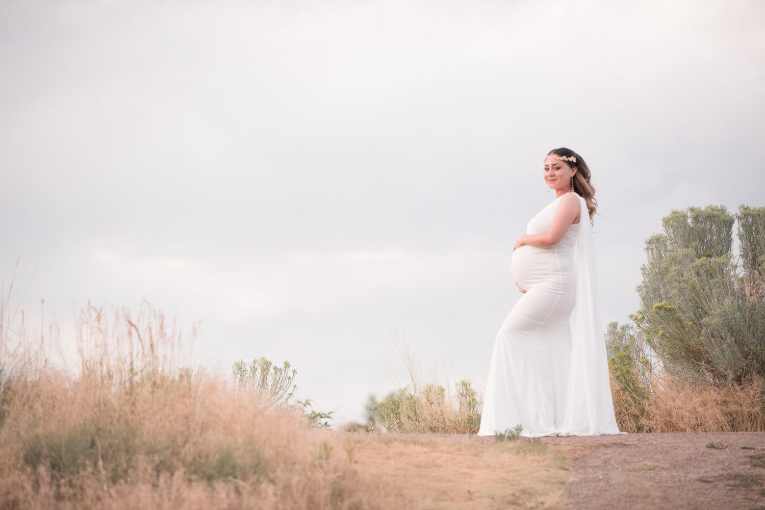 Pregnant woman in flowing white dress stands serenely in golden field under overcast sky.