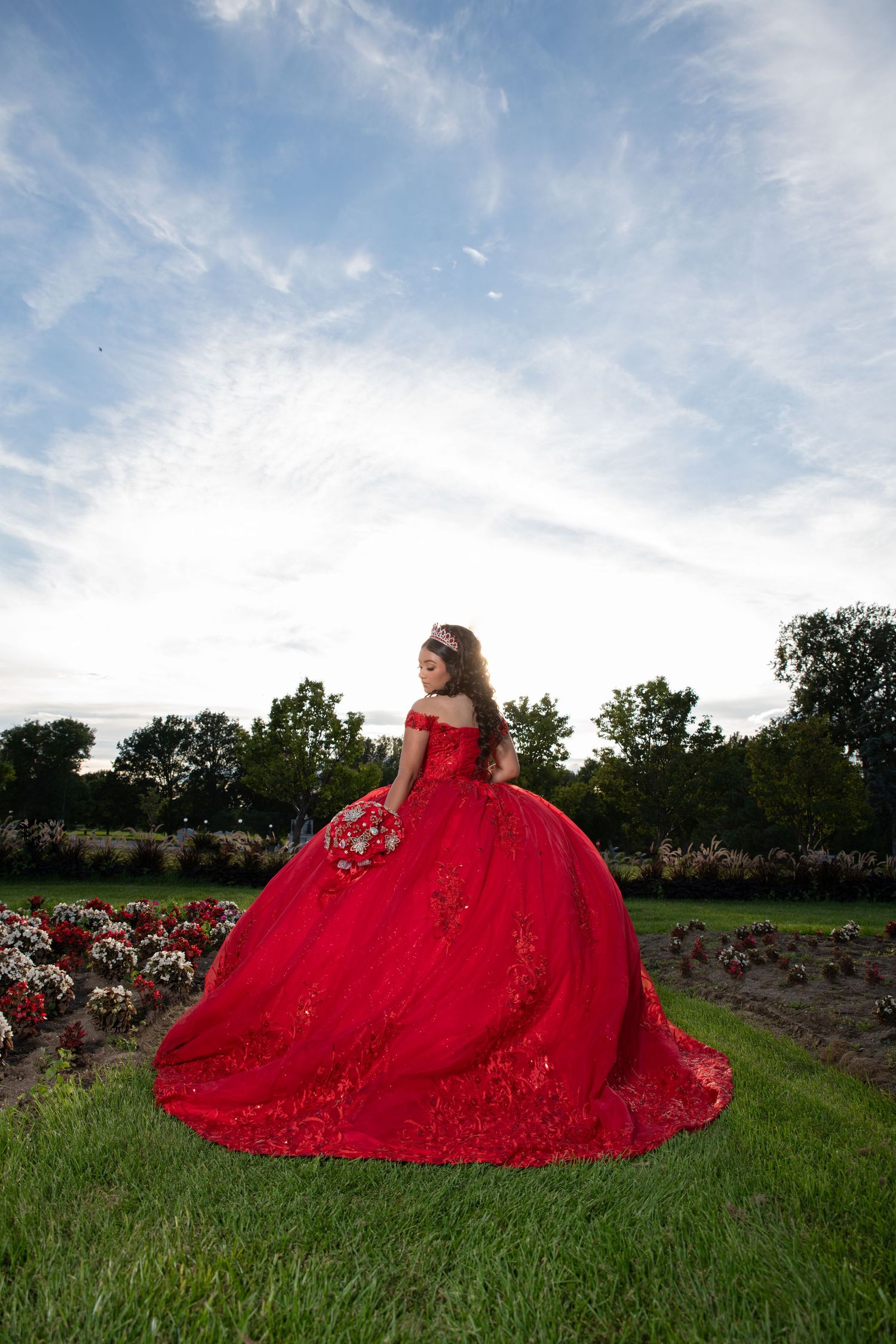 Young woman in red off-shoulder quinceañera gown with long train stands in garden under cloudy sky.