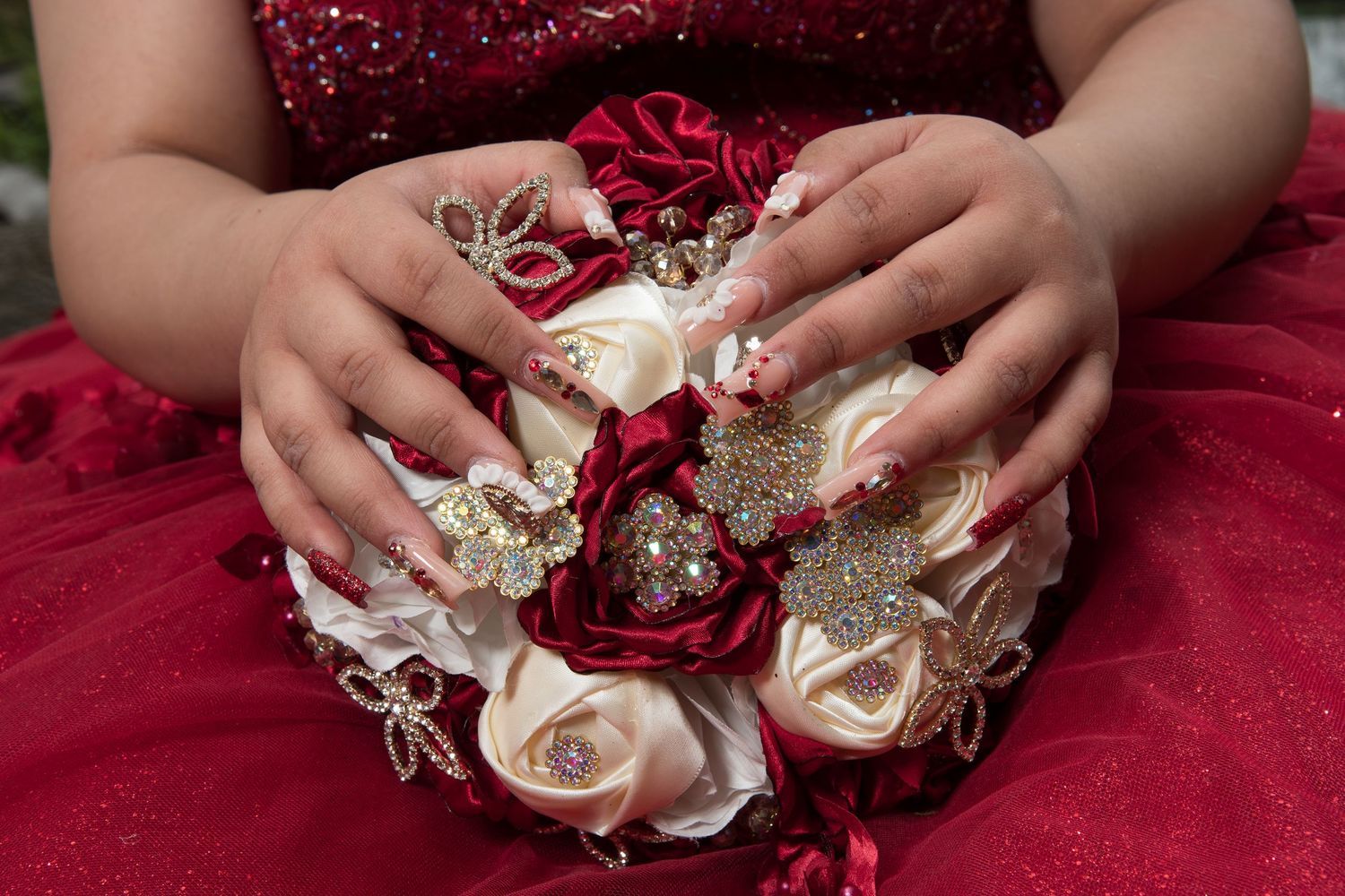 Close-up of manicured hands with rhinestone nail art holding ornate red and white jeweled bouquet.