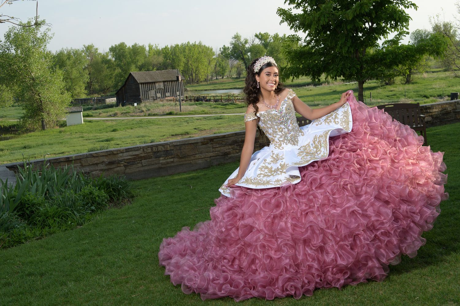 Young woman in pink and white quinceañera ball gown sits gracefully on green garden steps.