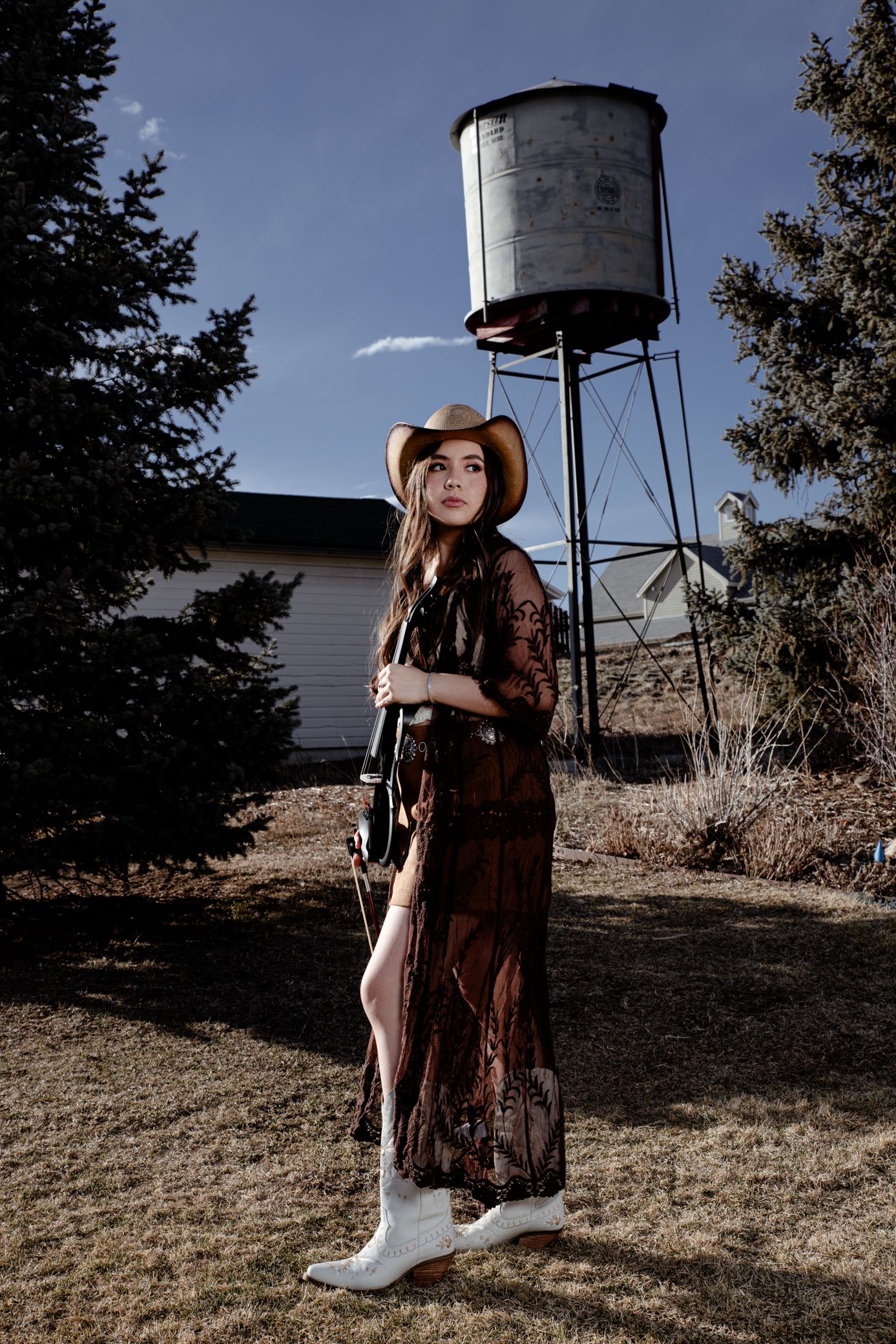 Young woman in cowboy hat, lace dress, and white boots standing near water tower and pine trees.