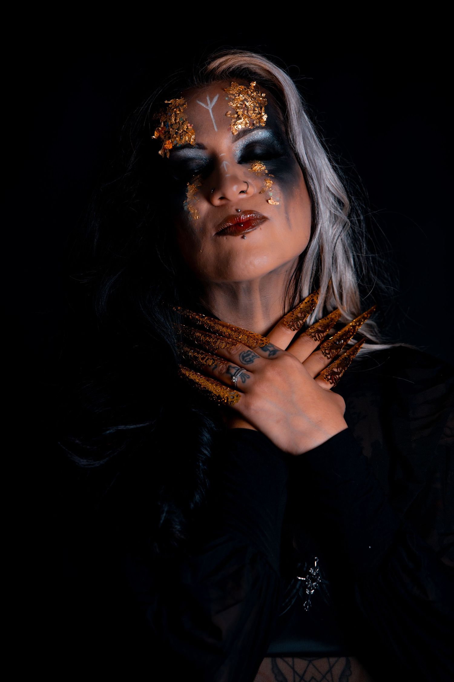 Woman with gold foil eye makeup and long gilded nails poses dramatically against black background.