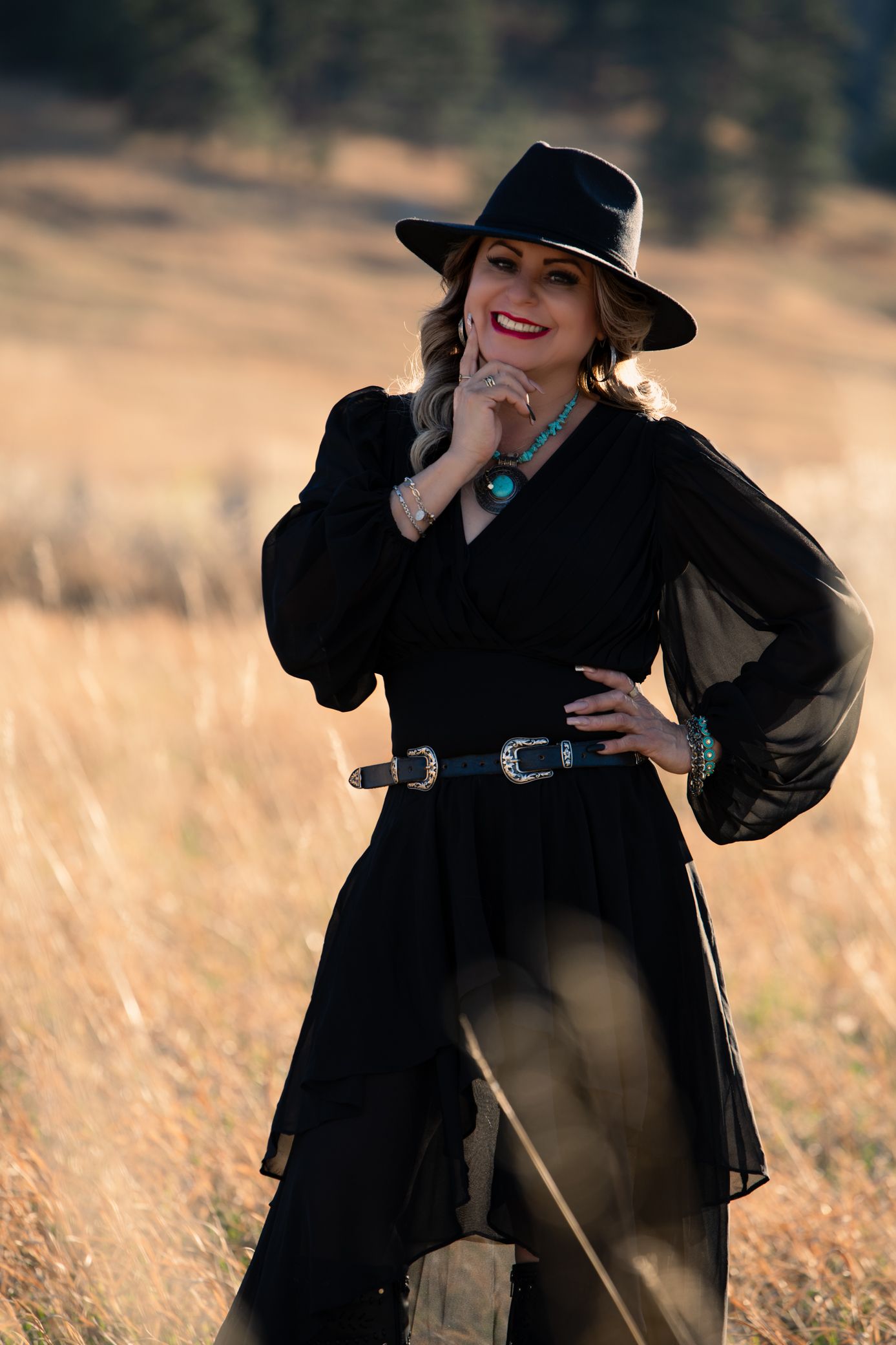 Smiling blonde woman in black dress with western belt and hat posing in golden autumn meadow.