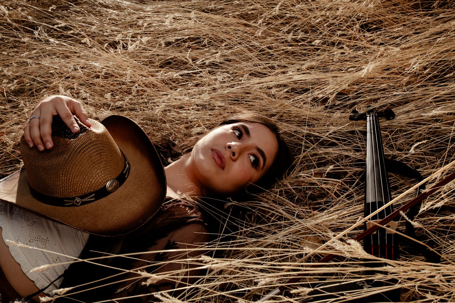 Woman lying in wheat field holding cowboy hat with violin beside her in warm sunlight.