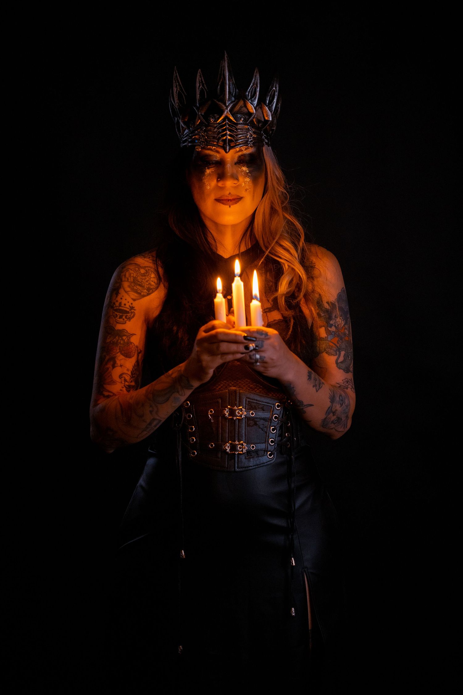 Tattooed woman wearing metal crown holds lit candles in dark atmospheric portrait photography.