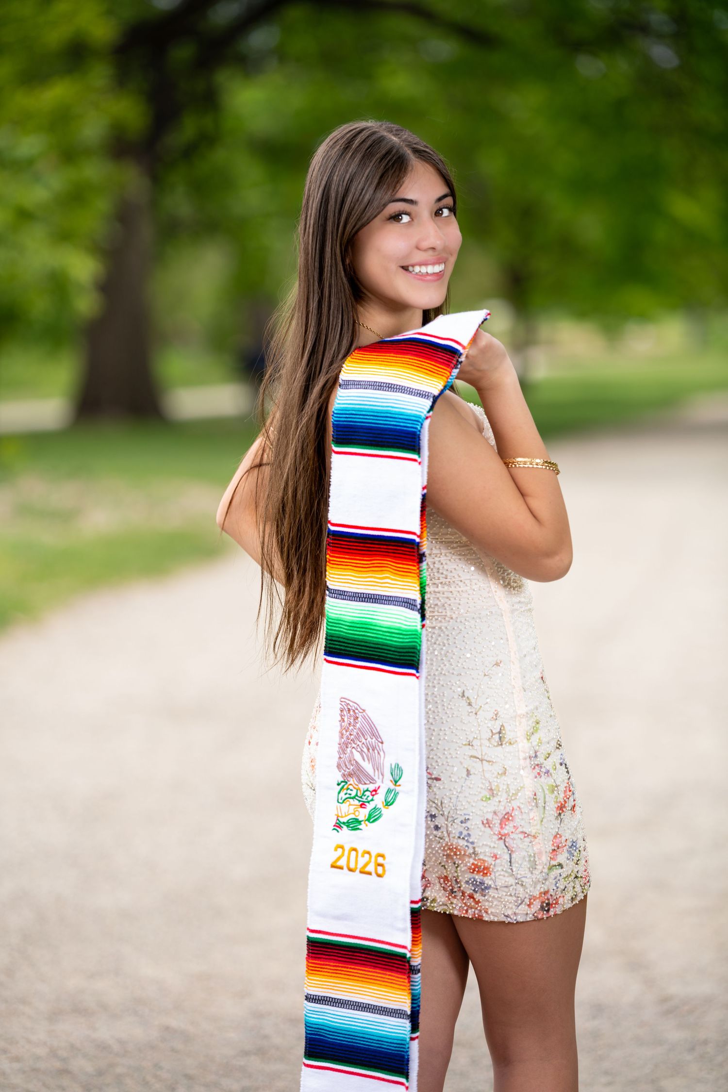 Smiling young woman holding colorful Mexican graduation stole with Class of 2026 in sunny park setting.