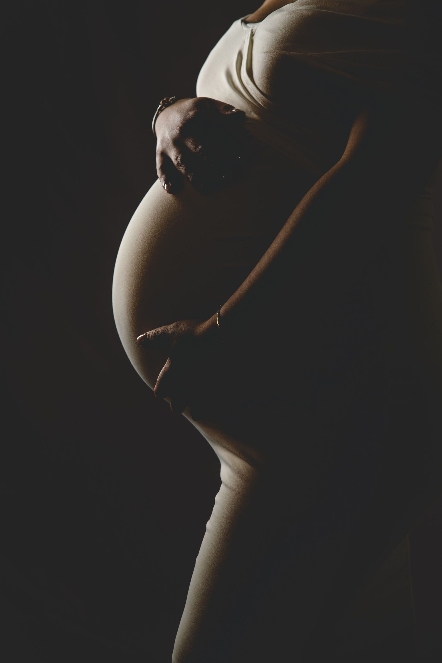 Dramatic silhouette of pregnant woman cradling baby bump against dark black background.