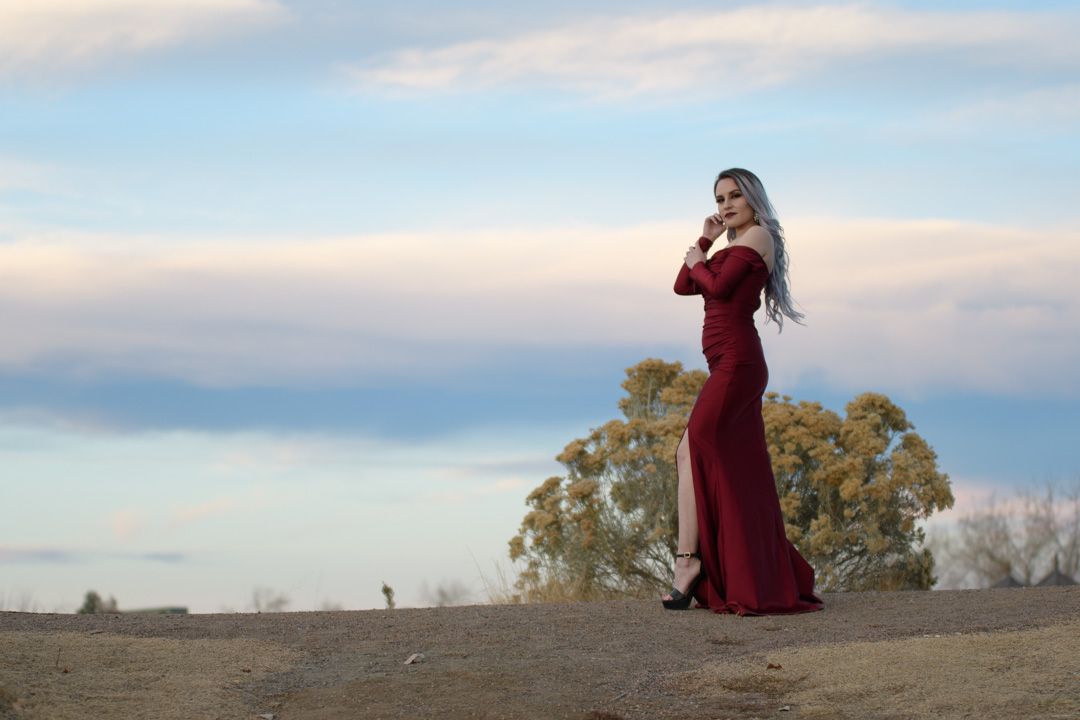 A woman in an elegant red gown poses on sandy terrain with trees and cloudy sky backdrop.
