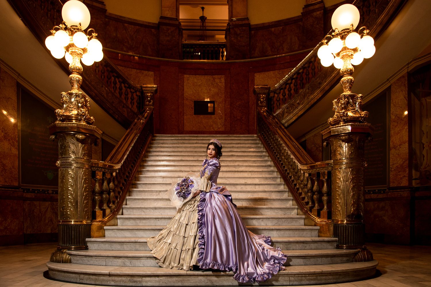 Woman in lavender and gold Victorian ball gown posing on grand ornate staircase with chandeliers.