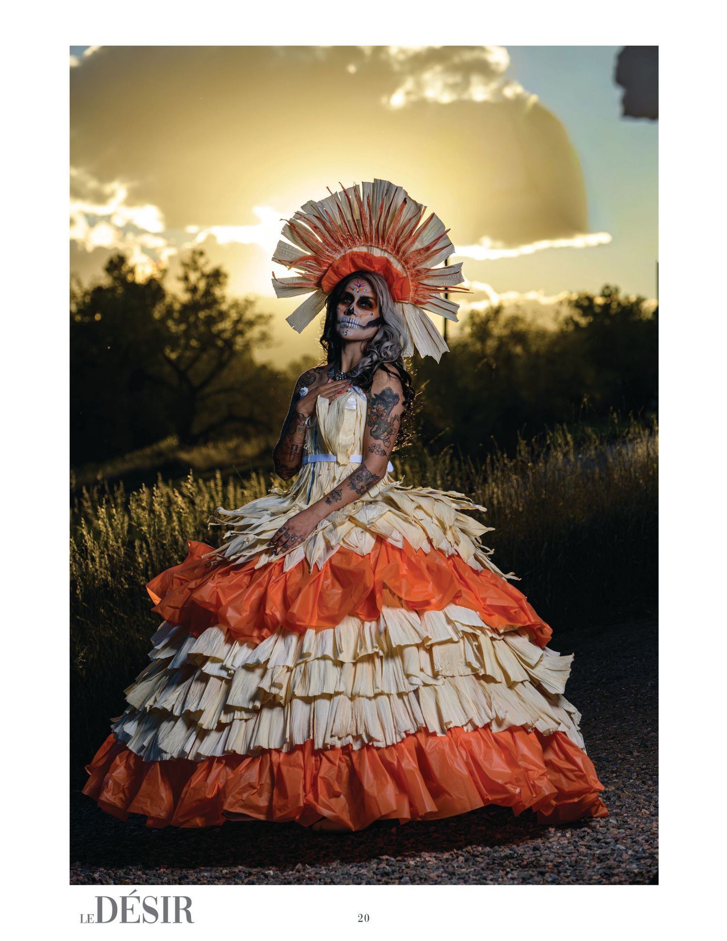 Woman in skull face paint and a radiating headdress wears a tiered orange and cream ruffled gown in a field at golden-hour sunset.