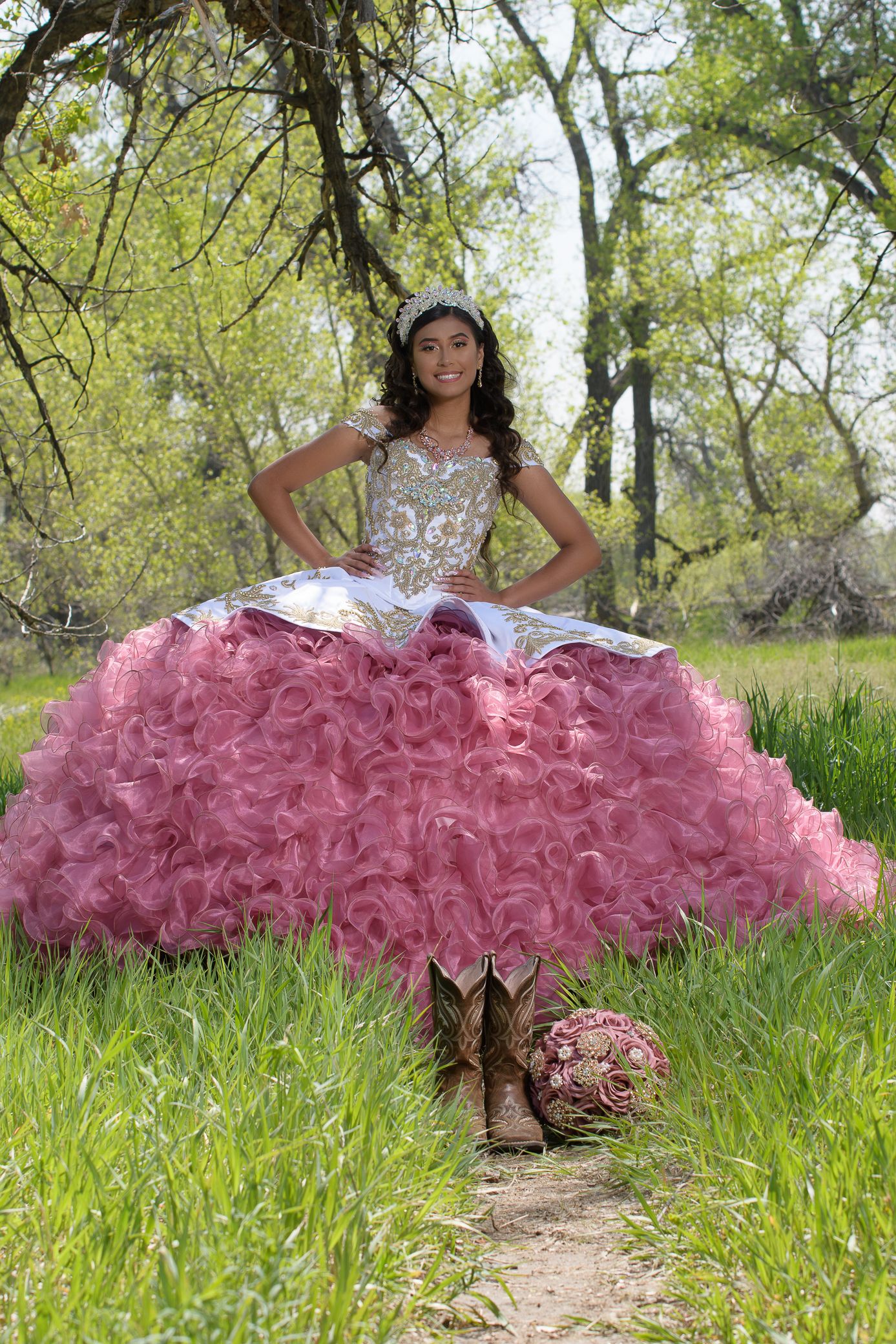 Young woman in pink ruffled quinceañera ball gown with gold bodice poses in green meadow.