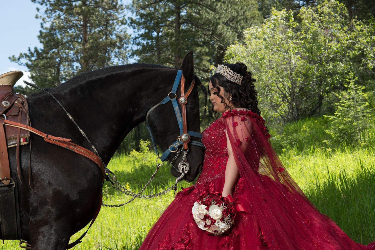 Young woman in red quinceañera gown with tiara holds bouquet beside black horse in lush green forest.