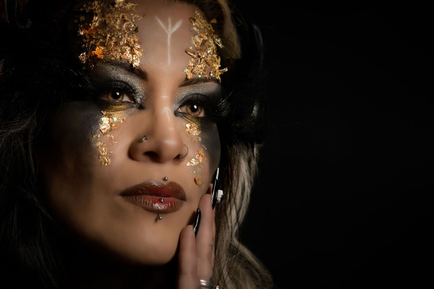 Close-up portrait of woman with elaborate gold glitter and black fantasy makeup on dark background.