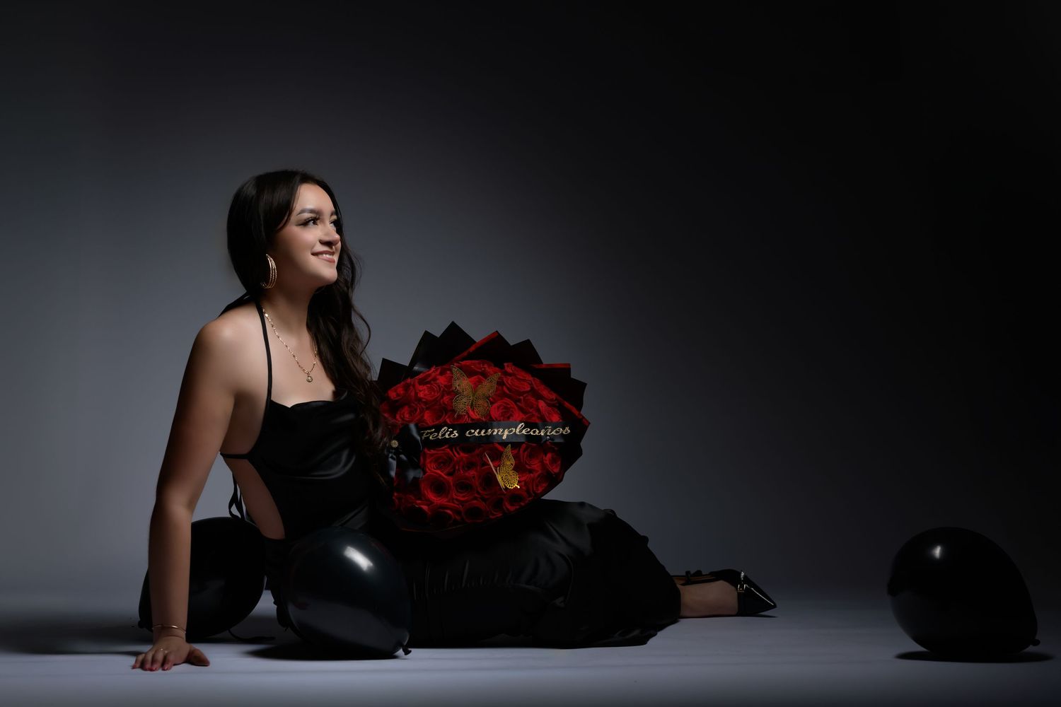 Woman in black outfit sitting on floor holding red rose bouquet with black balloons in dark studio setting. Birthday photoshoot