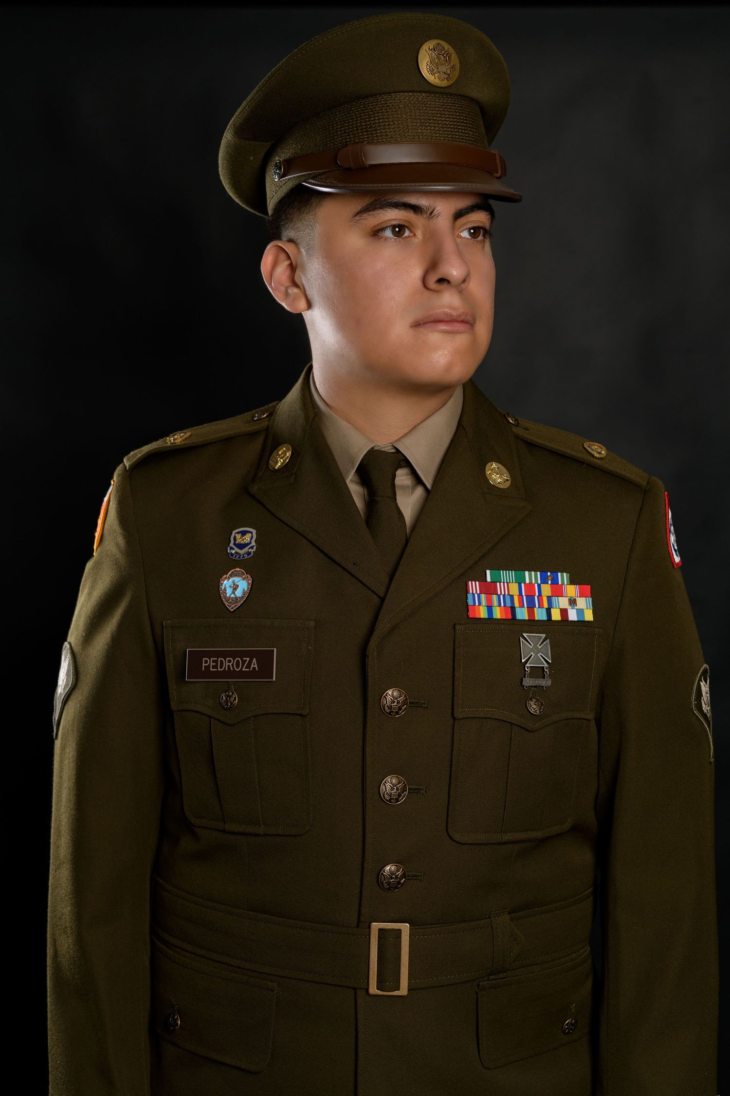 Young US Army soldier in formal dress uniform with medals and ribbons on dark background.