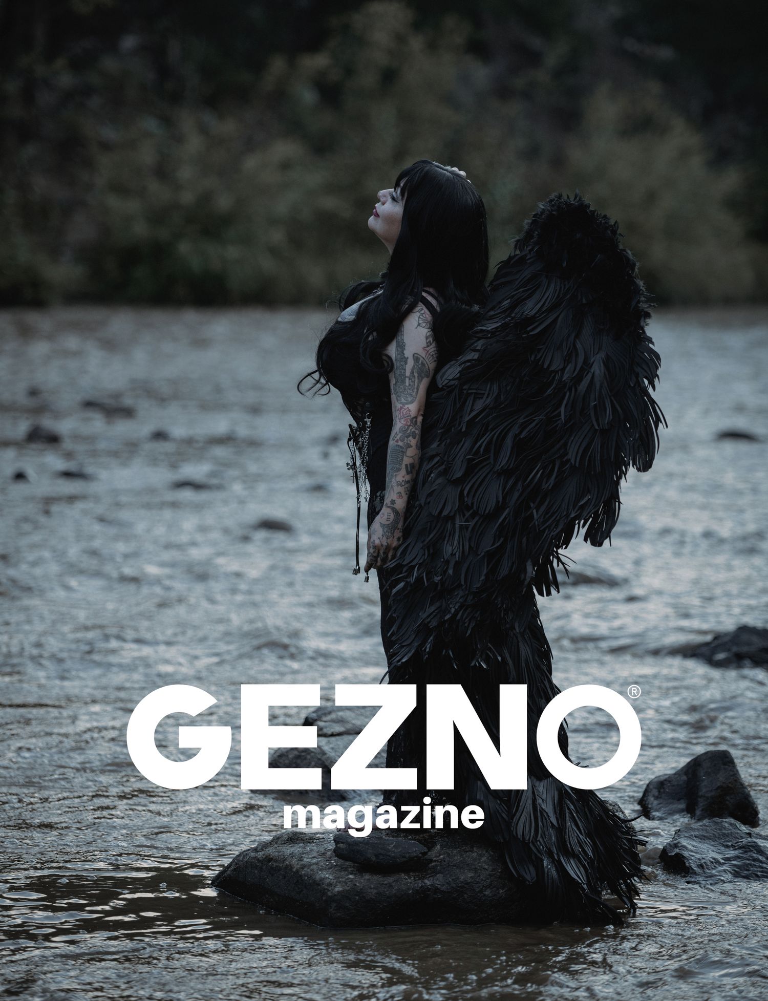 Tattooed woman with long black hair and large black feathered wings stands on a rock in a shallow river on a Gezno Magazine cover.