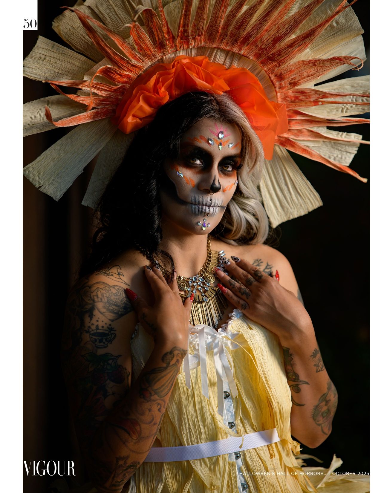 Woman in skull face paint with gems, a sunburst corn husk headdress, tattoos and a yellow dress in a Day of the Dead style portrait.