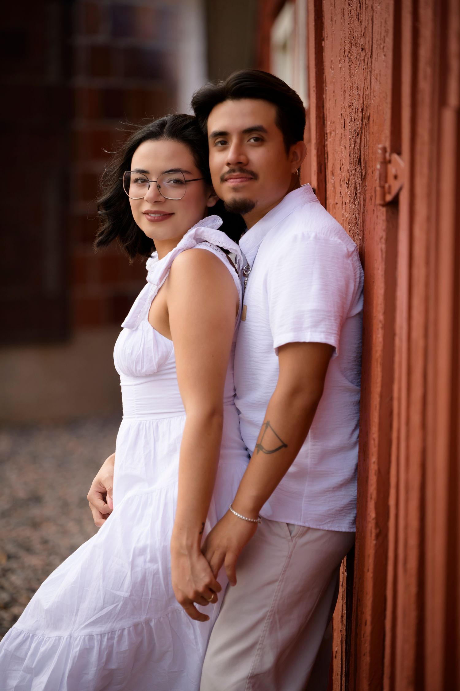 A young couple in matching white outfits hold hands while posing against a rustic red wooden wall outdoors.