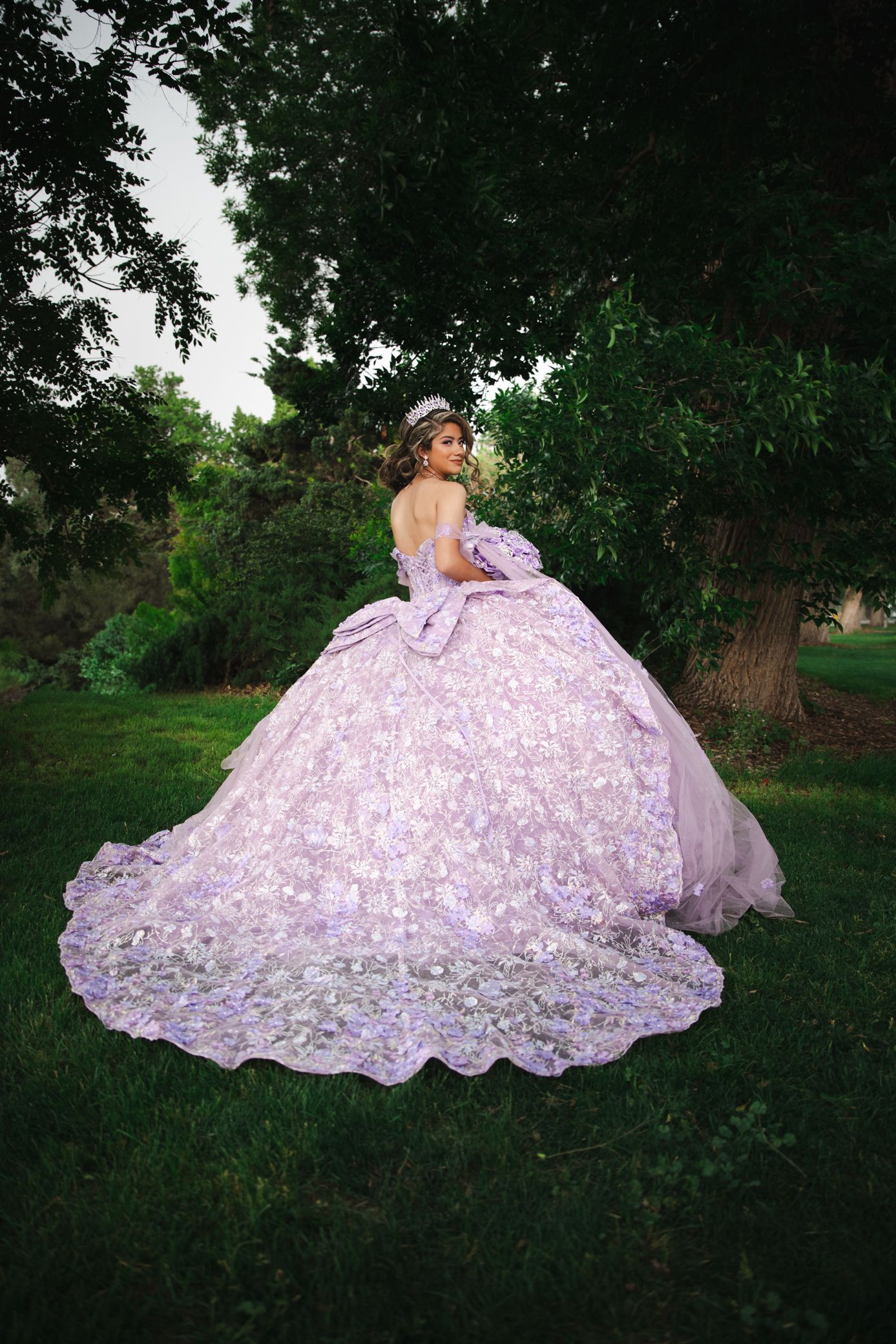 Quinceañera in stunning lavender ball gown with floral embroidery standing near trees outdoors.