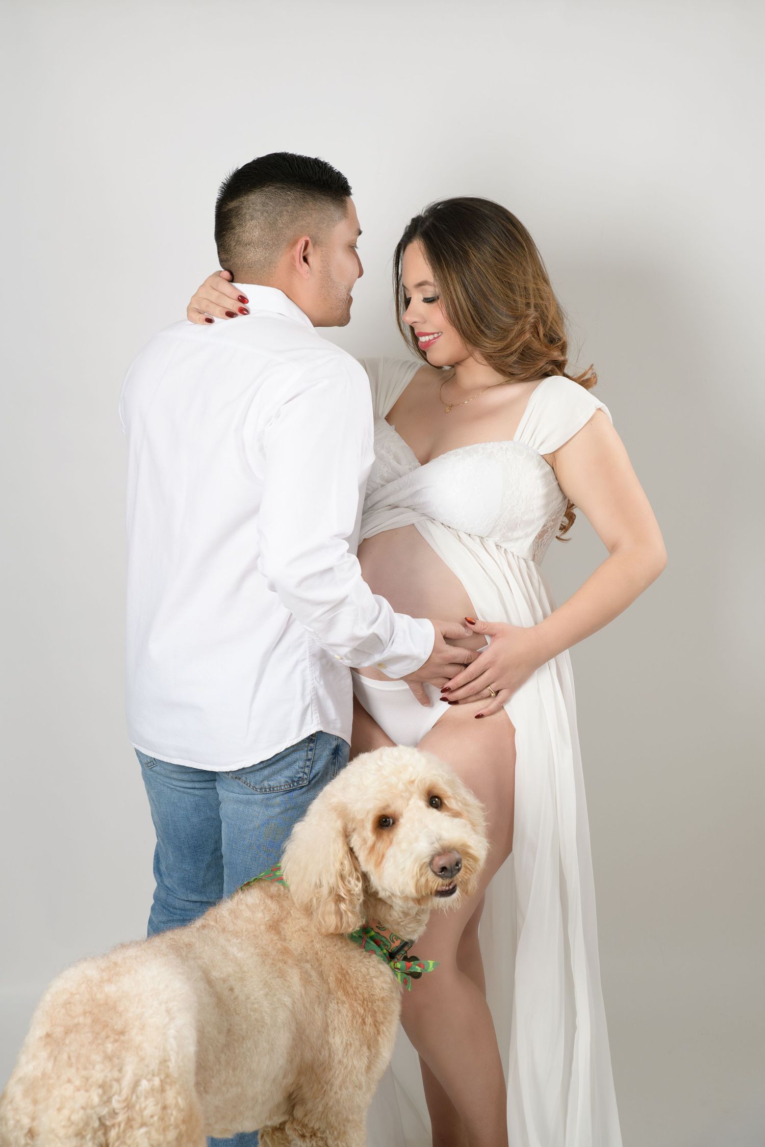 Expecting couple in white outfits share tender moment with golden doodle dog in studio.