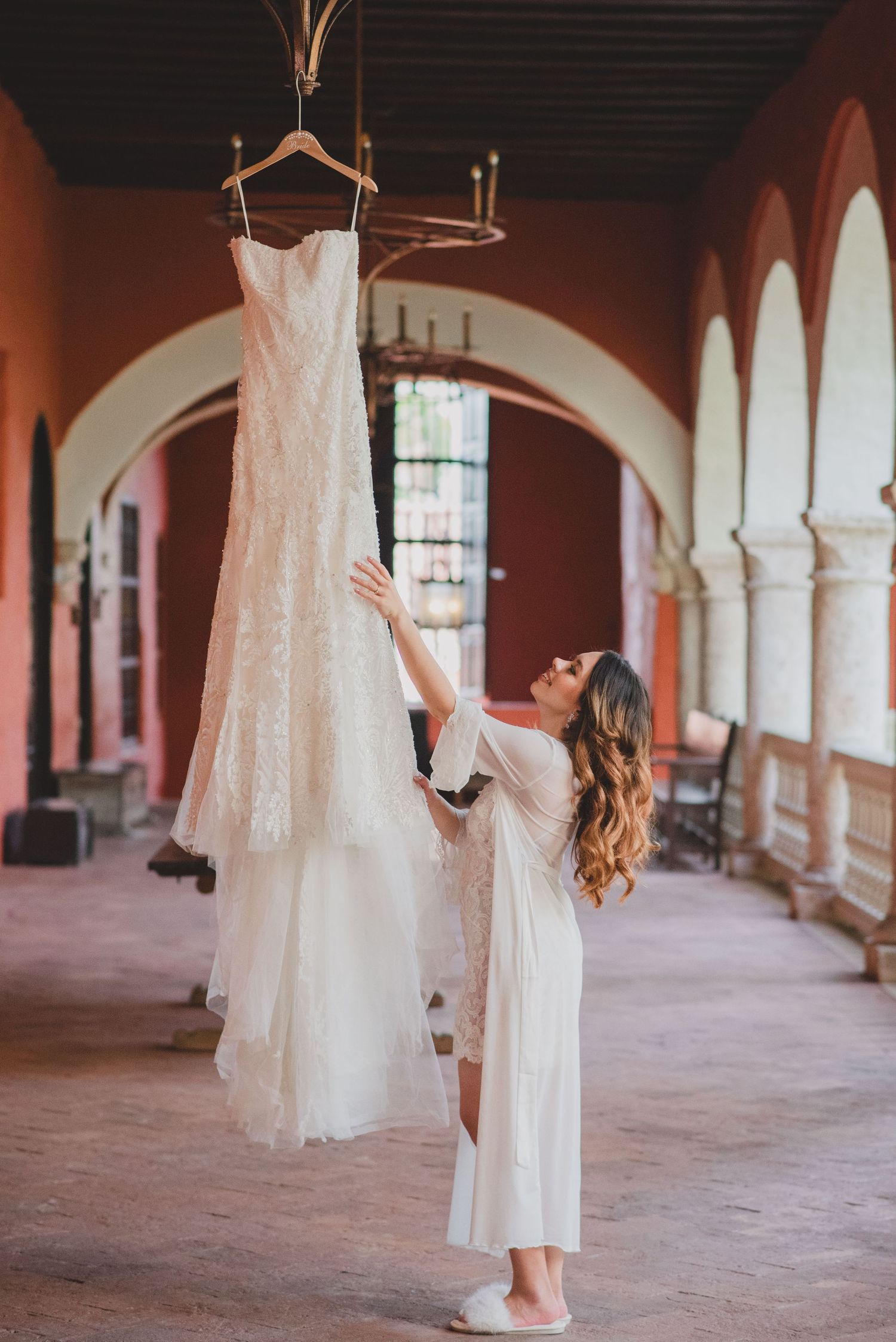 Bride in robe reaches toward lace wedding dress hanging in elegant arched colonial venue hallway.