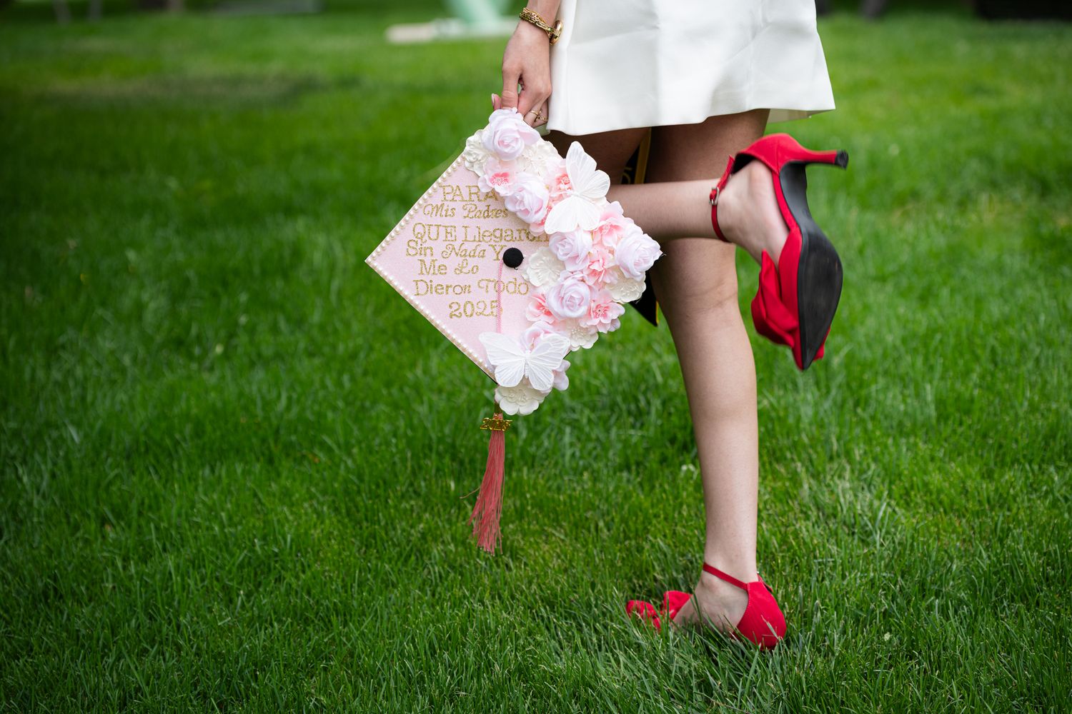 Graduate holding floral decorated cap beside red high heels while standing on lush green grass.