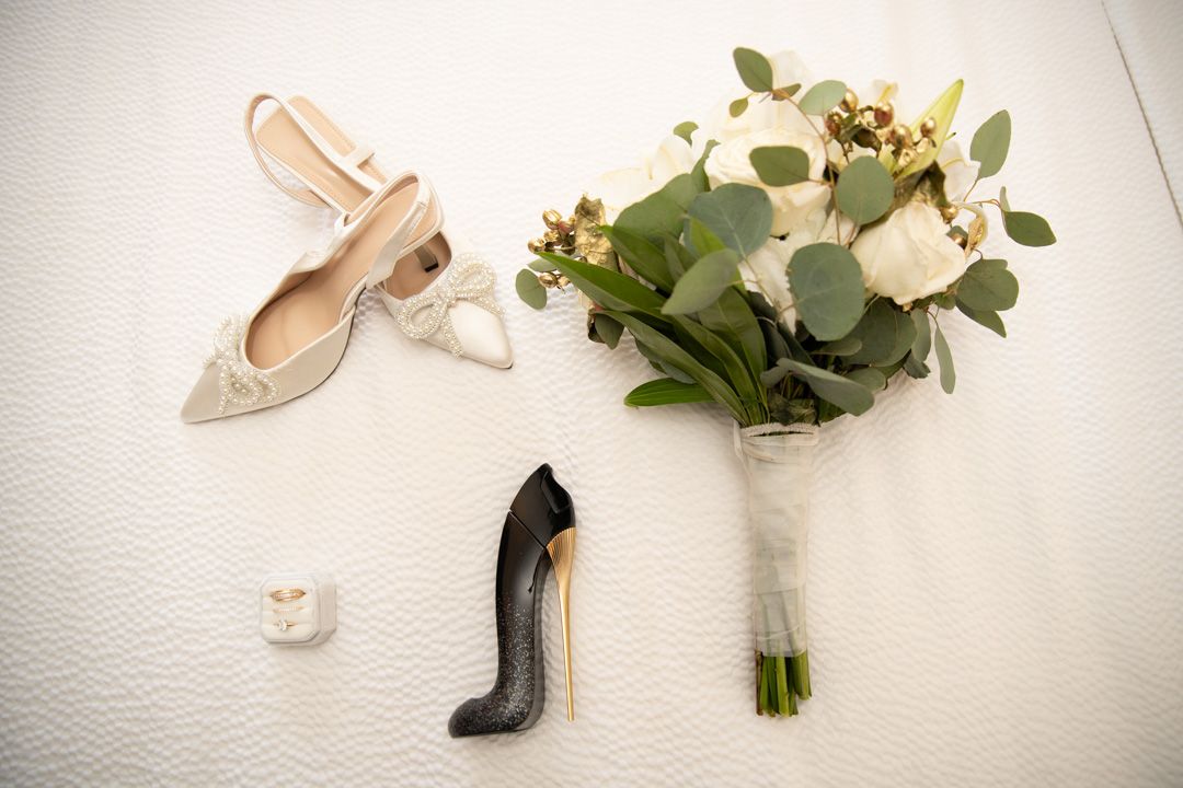 Bridal flat lay with nude heels, Carolina Herrera perfume, and white floral bouquet on white surface.