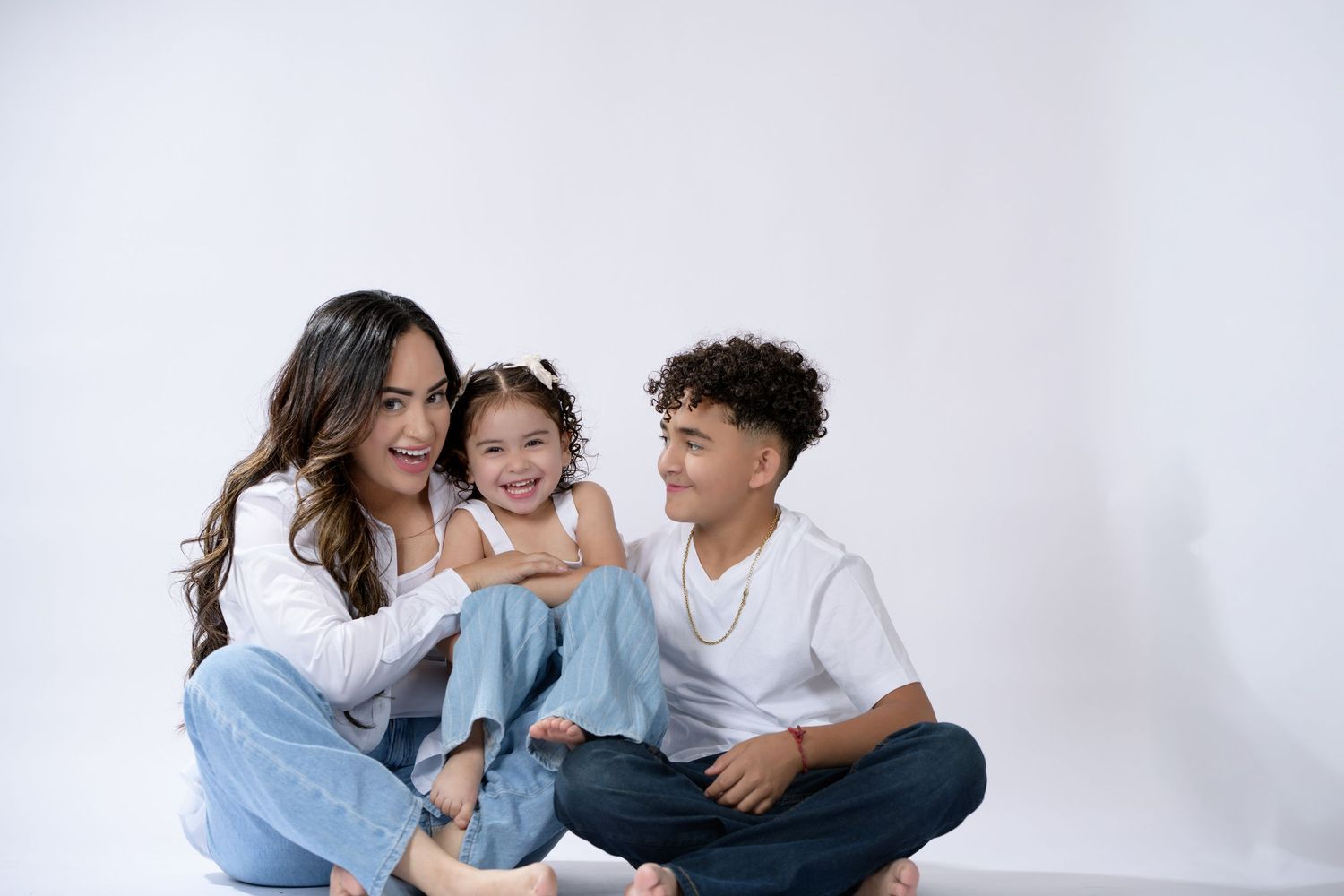 A woman and two children sit together smiling on a white background, wearing casual jeans and white tops.