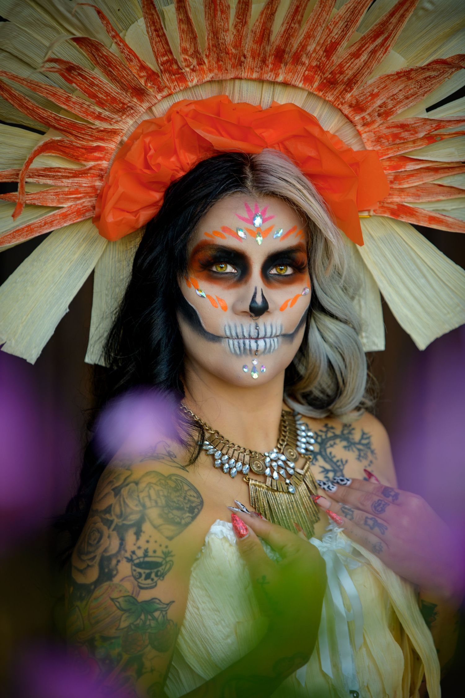 Colorful Day of the Dead sugar skull makeup with orange floral crown and tribal necklace on tattooed woman.