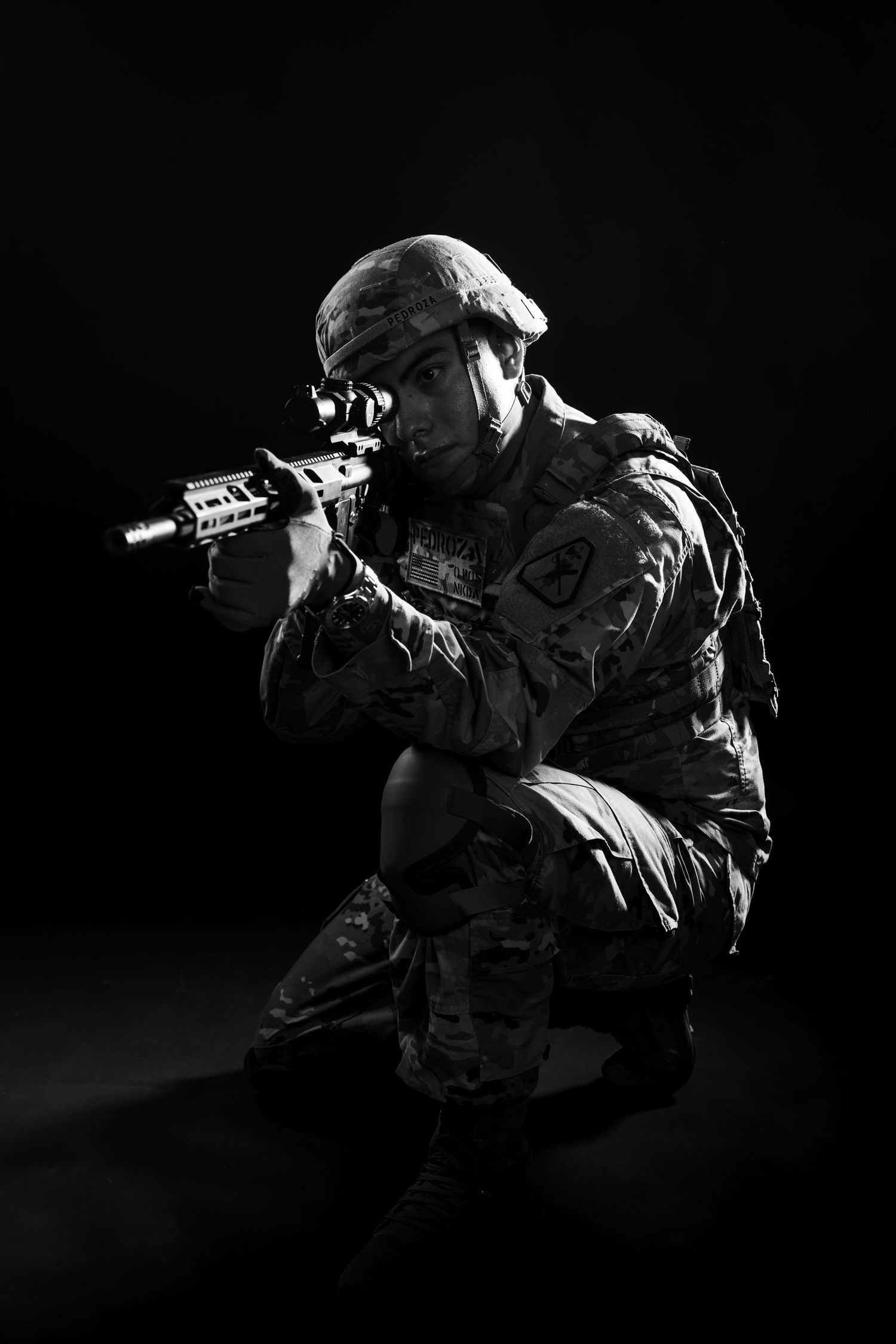 Black and white photo of US Army soldier in combat gear aiming rifle on dark background.