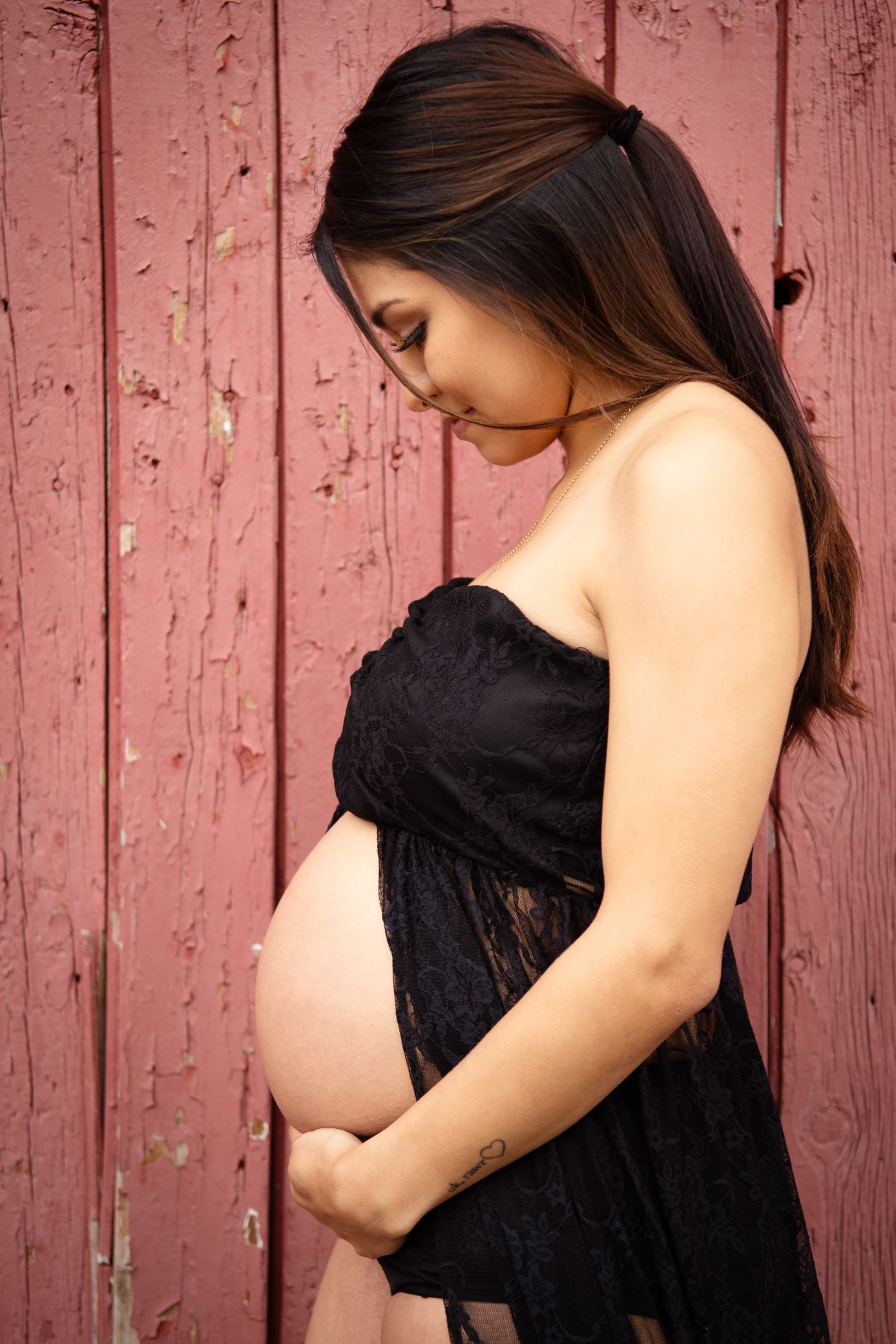 Pregnant woman in black lace dress cradling baby bump against rustic red wooden barn wall.