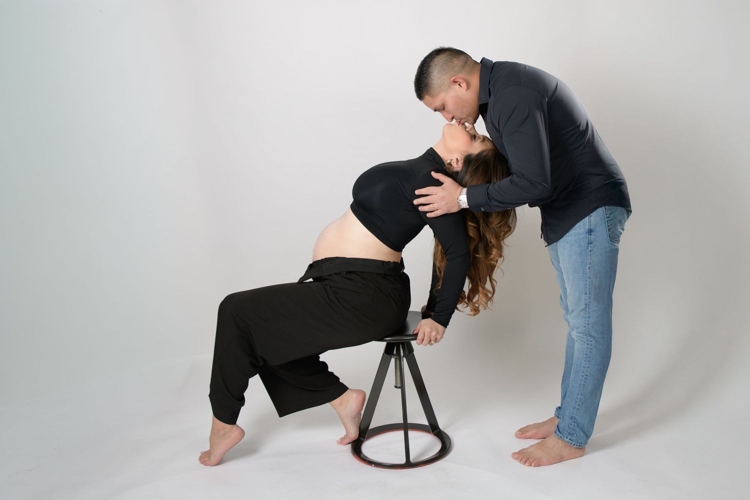 Couple shares tender kiss during maternity photoshoot, woman seated on stool in studio setting.