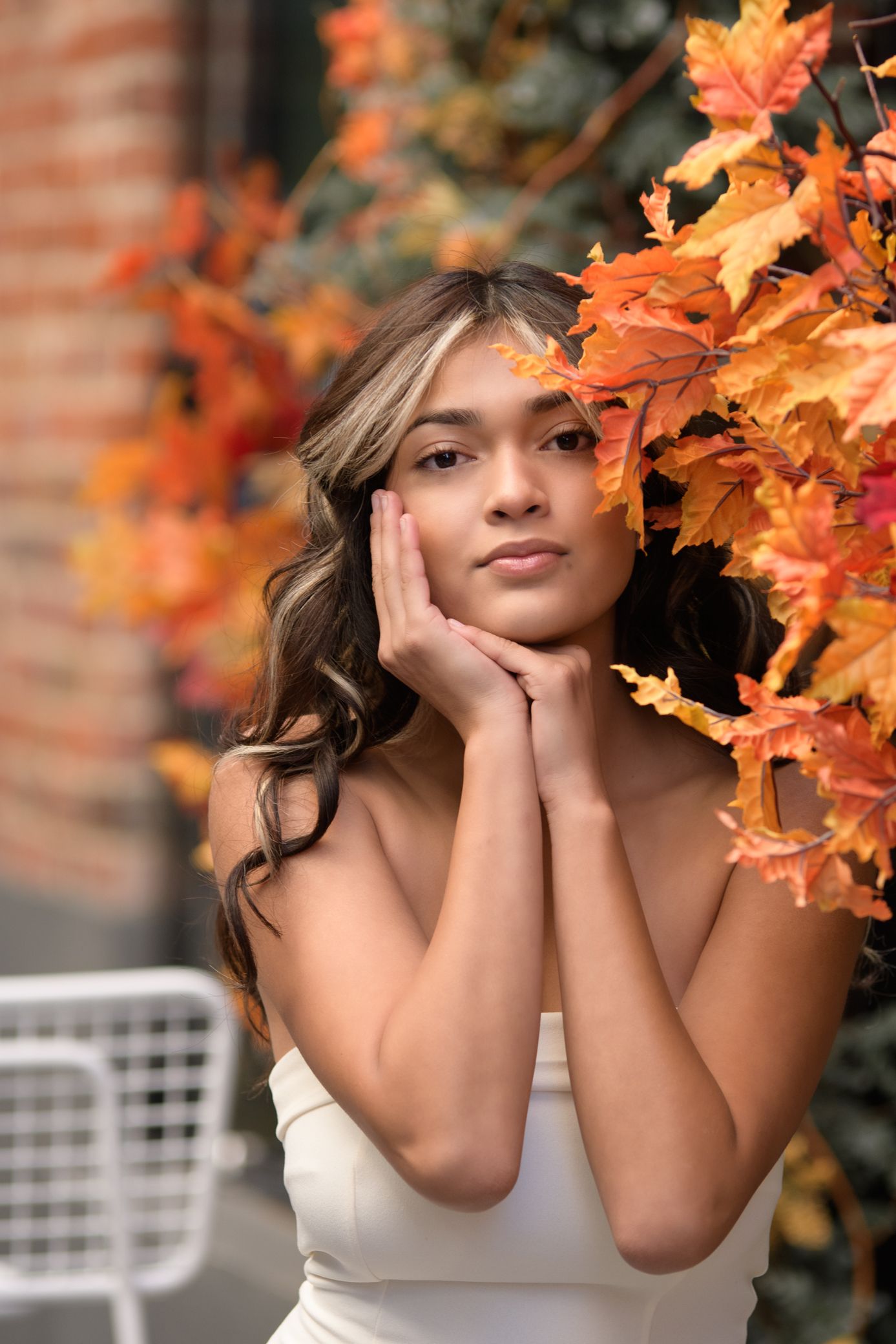 Young woman in white dress posing with hands on cheeks beside vibrant orange autumn maple leaves.