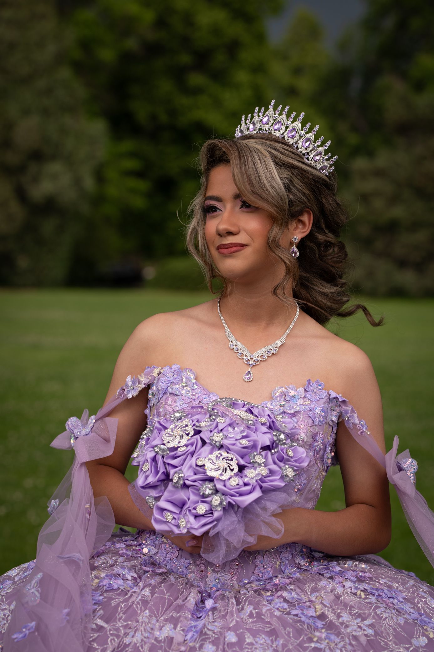 Young woman in lavender quinceañera ball gown with tiara holding purple flower bouquet.