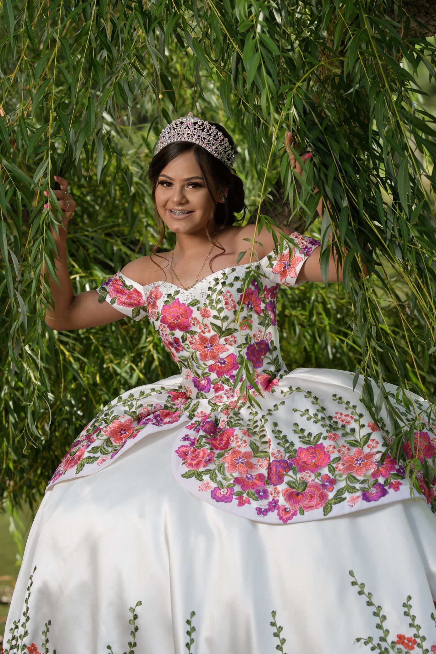 Smiling quinceañera in white off-shoulder floral ball gown with tiara under willow tree.