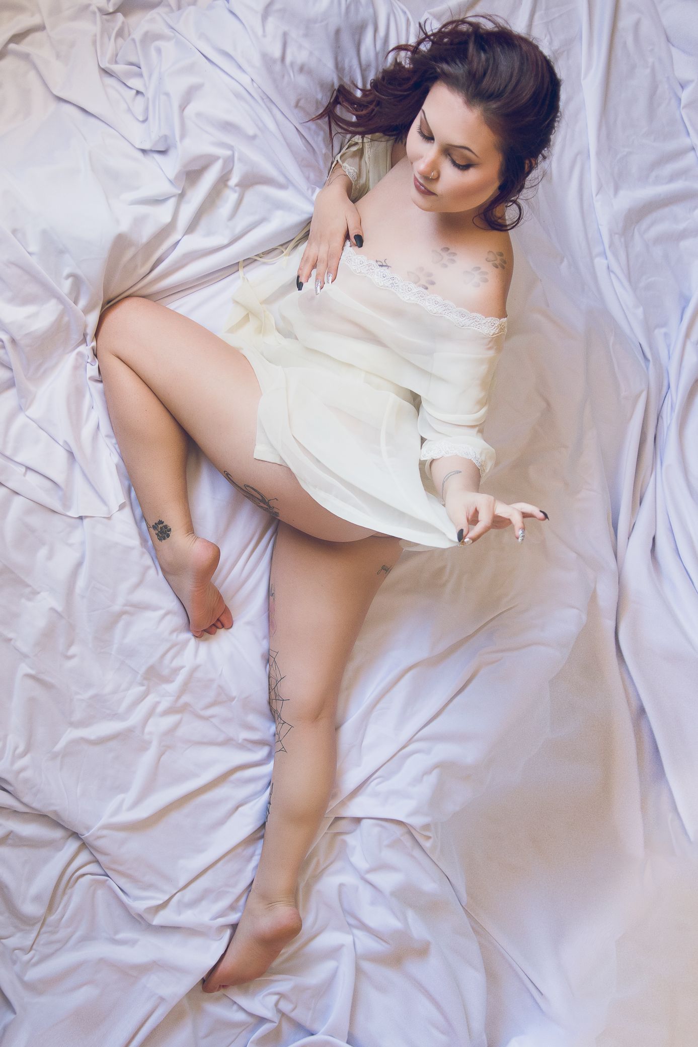 A tattooed brunette woman in white sheer robe reclines gracefully on white satin bedding.