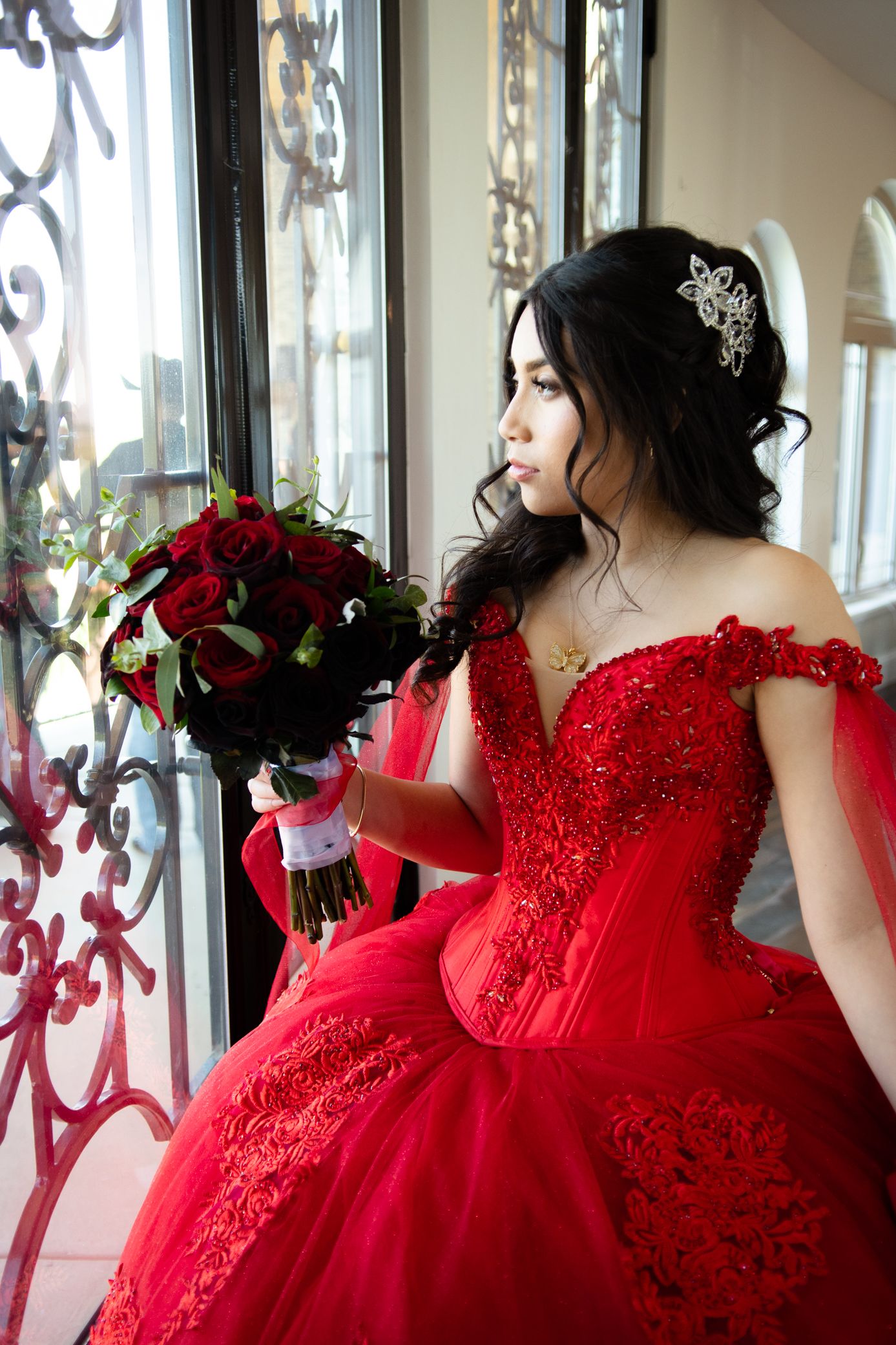 Young woman in red lace quinceañera gown gazes out window holding dark red rose bouquet.