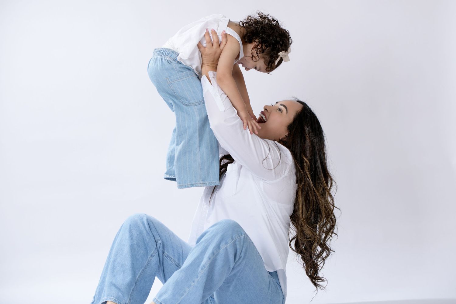 A joyful mother lifts her curly-haired toddler overhead while sitting on a white background, both wearing blue jeans.