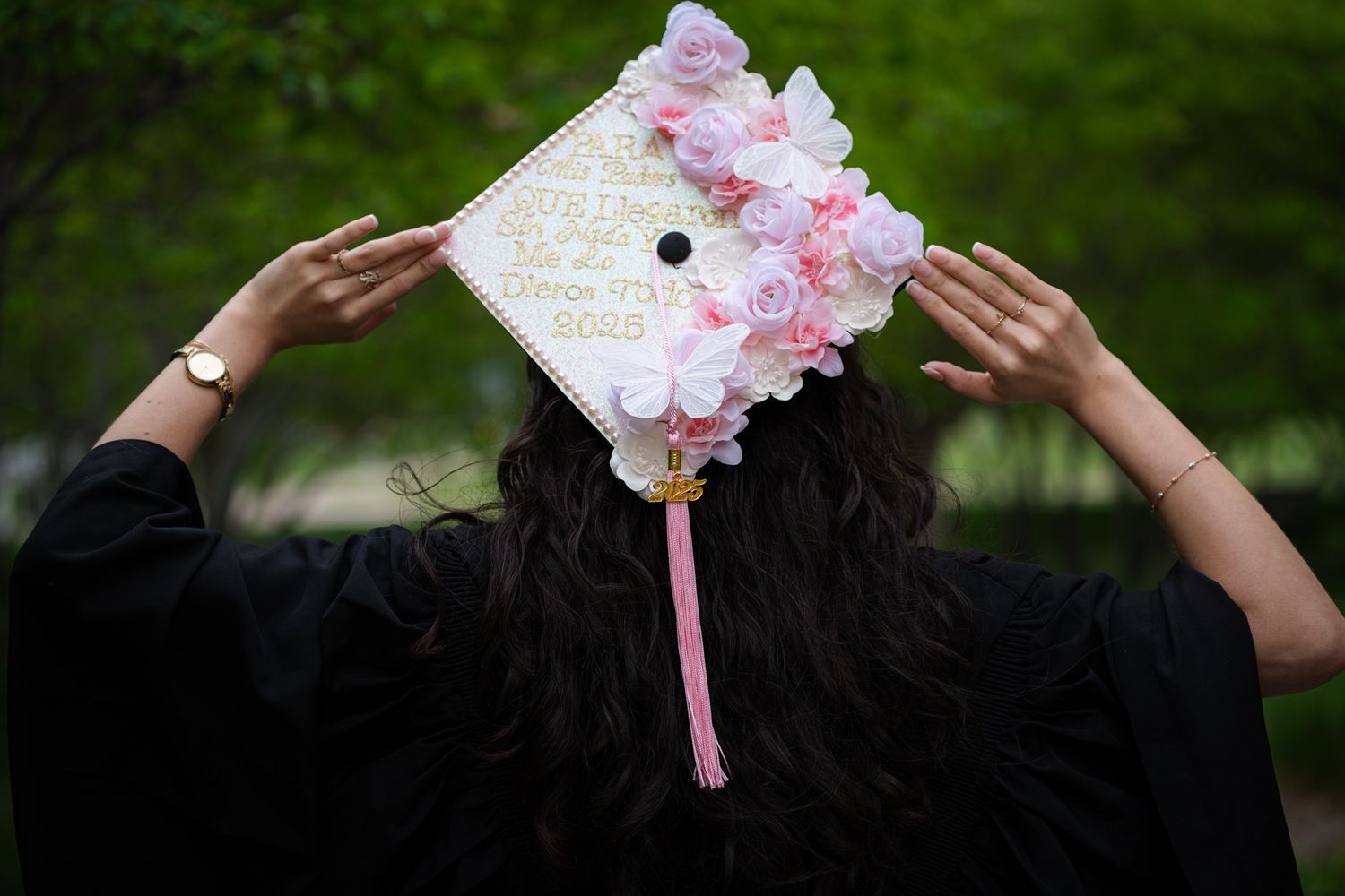 Graduate holding decorated pink floral graduation cap with 2025 message from behind in outdoor ceremony.