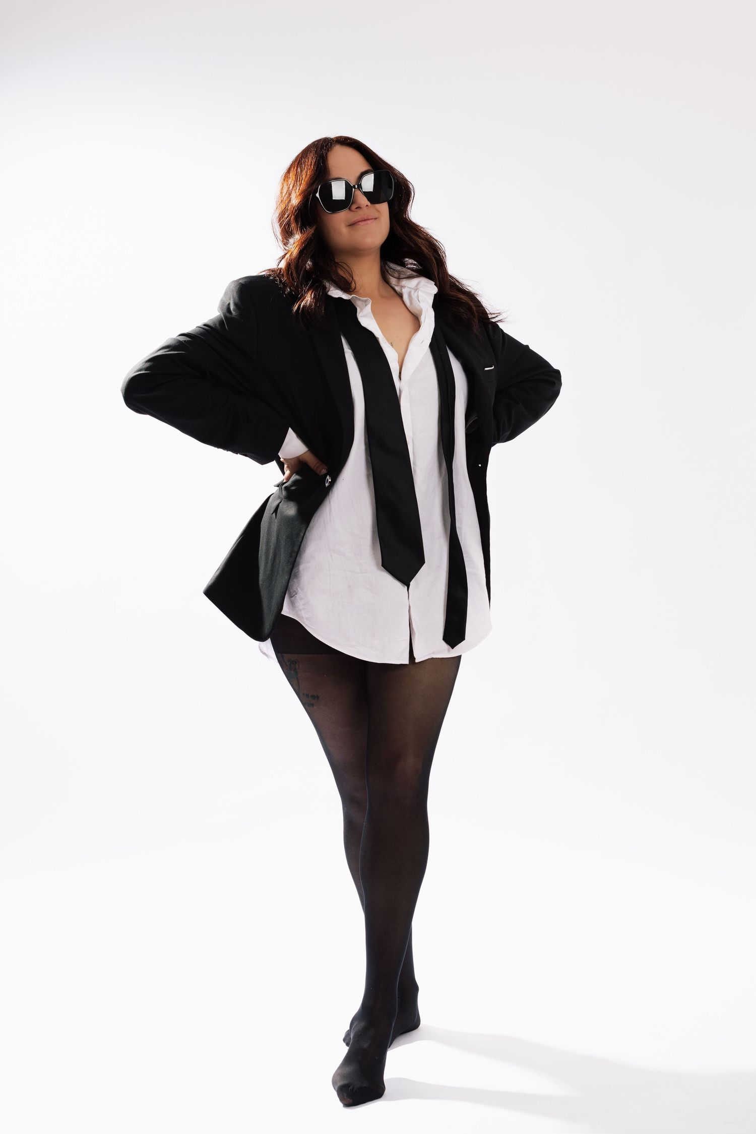 Woman in black blazer, white shirt, tie, tights, and heels poses confidently wearing sunglasses on white background.