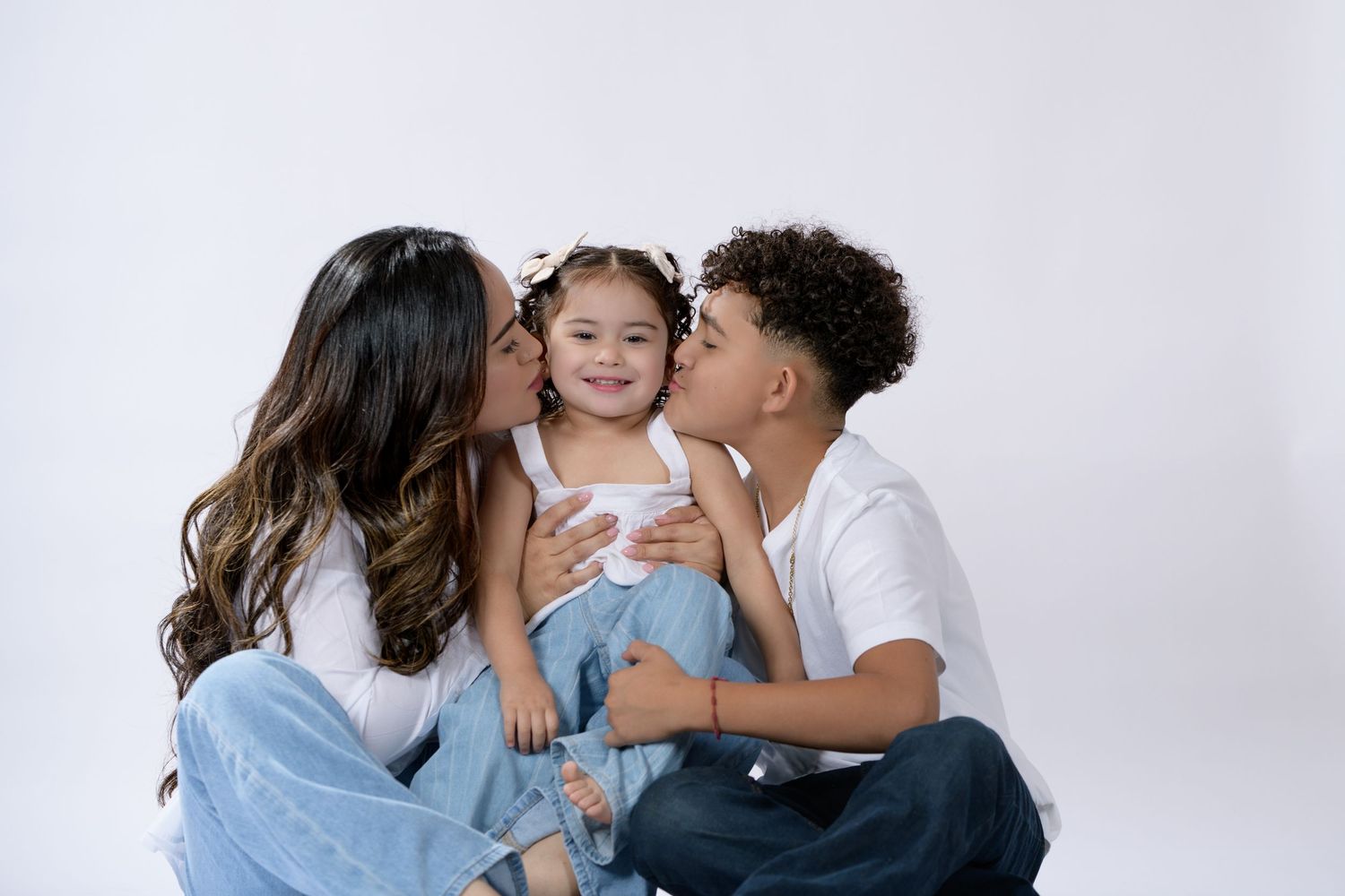 Two siblings kiss their smiling little sister on the cheeks in a sweet family portrait on a white background.