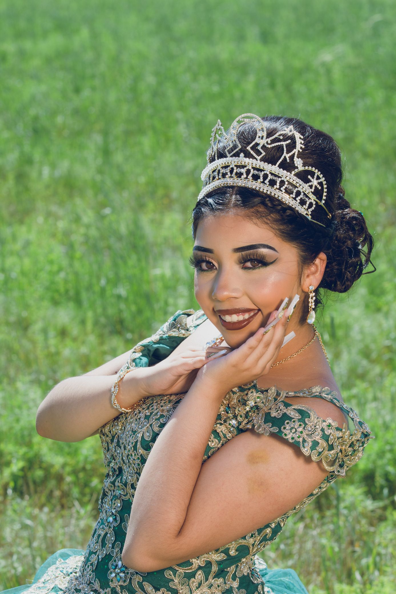 Joyful young woman in teal quinceañera dress and crystal tiara smiles while posing in green field.