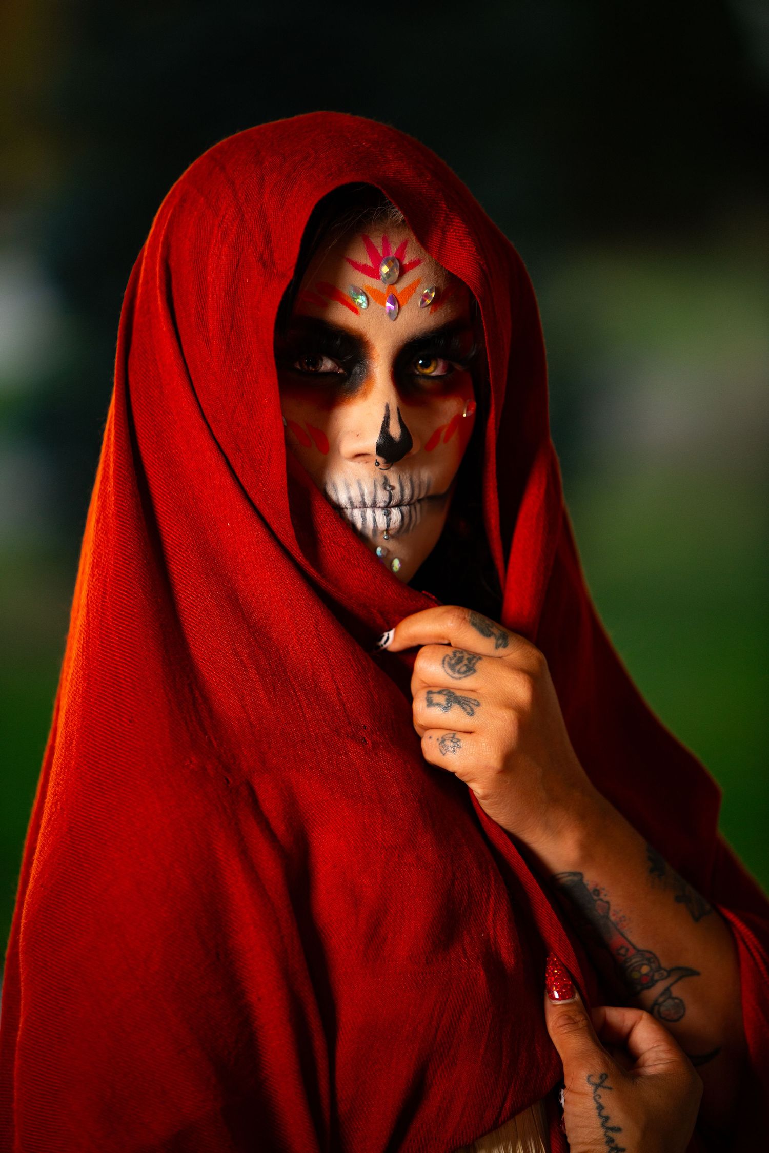 Woman in red hooded veil with colorful Day of the Dead skull makeup in outdoor portrait.
