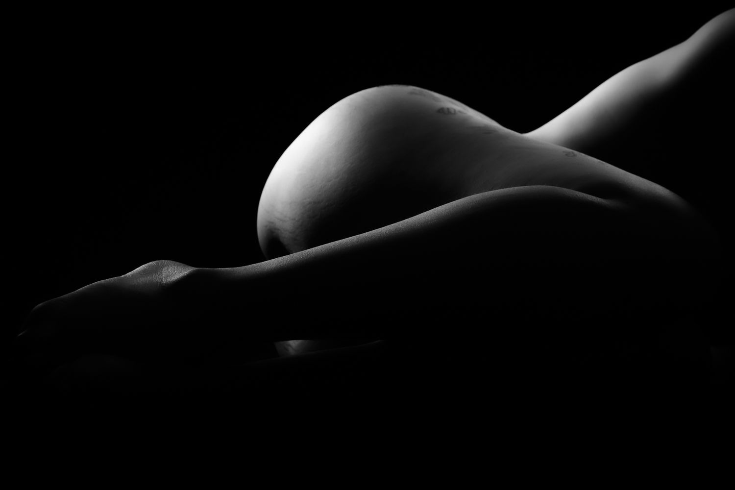 Artistic black and white bodyscape of pregnant woman lying down highlighting baby bump curves.