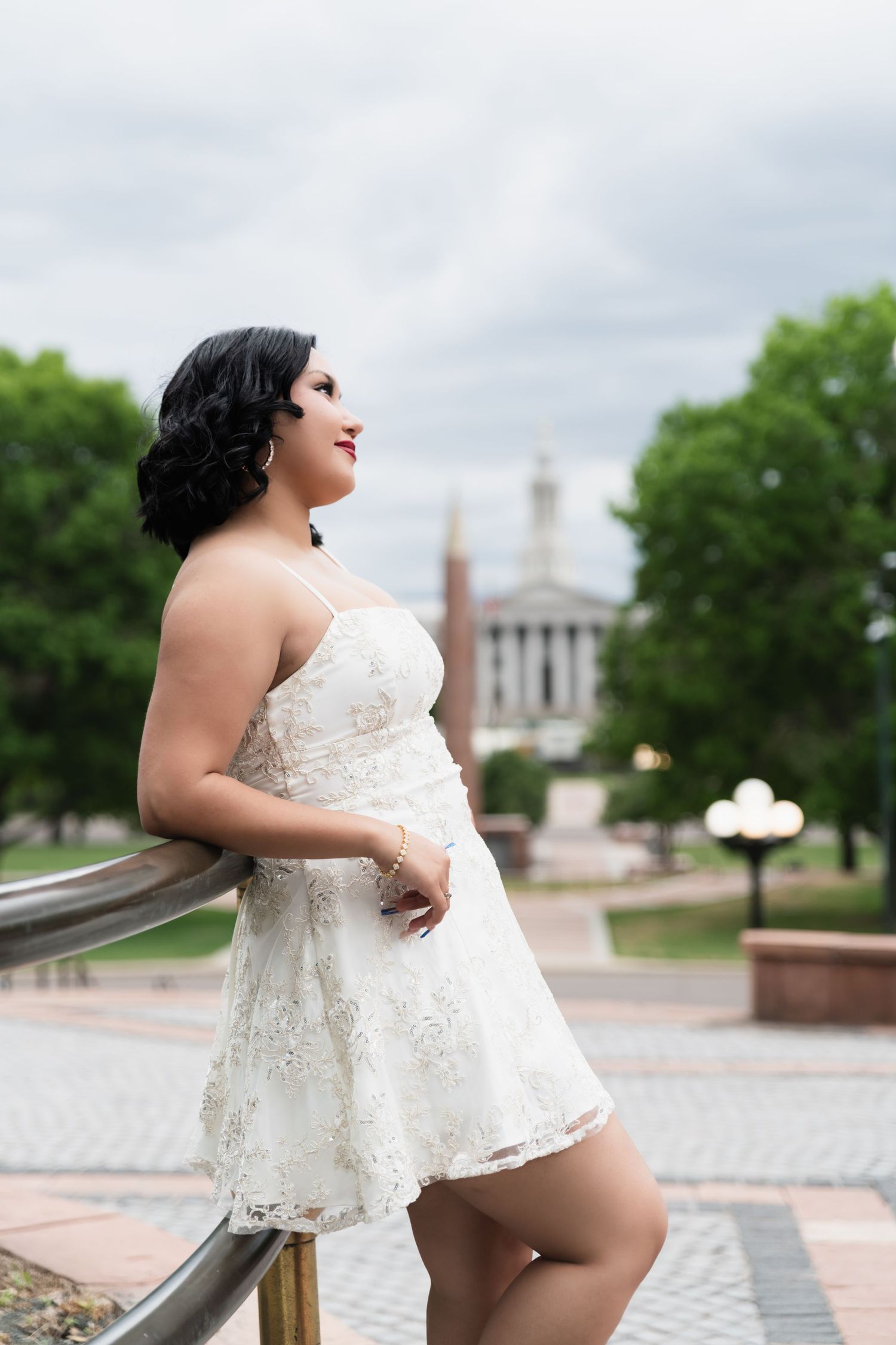 Elegant woman in white lace mini dress leans on railing with historic building in background.