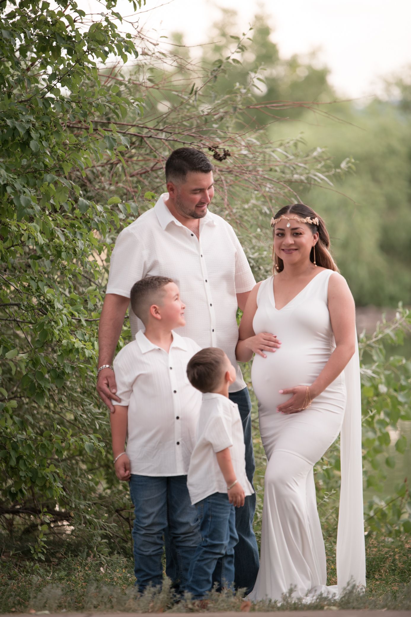 Pregnant woman in white dress with husband and two sons outdoors for a family maternity photo session.