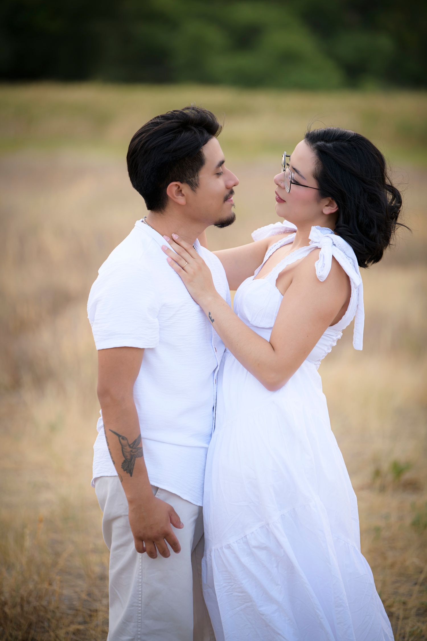Couple in white outfits embrace in a golden field, gazing lovingly at each other on a warm sunny day.