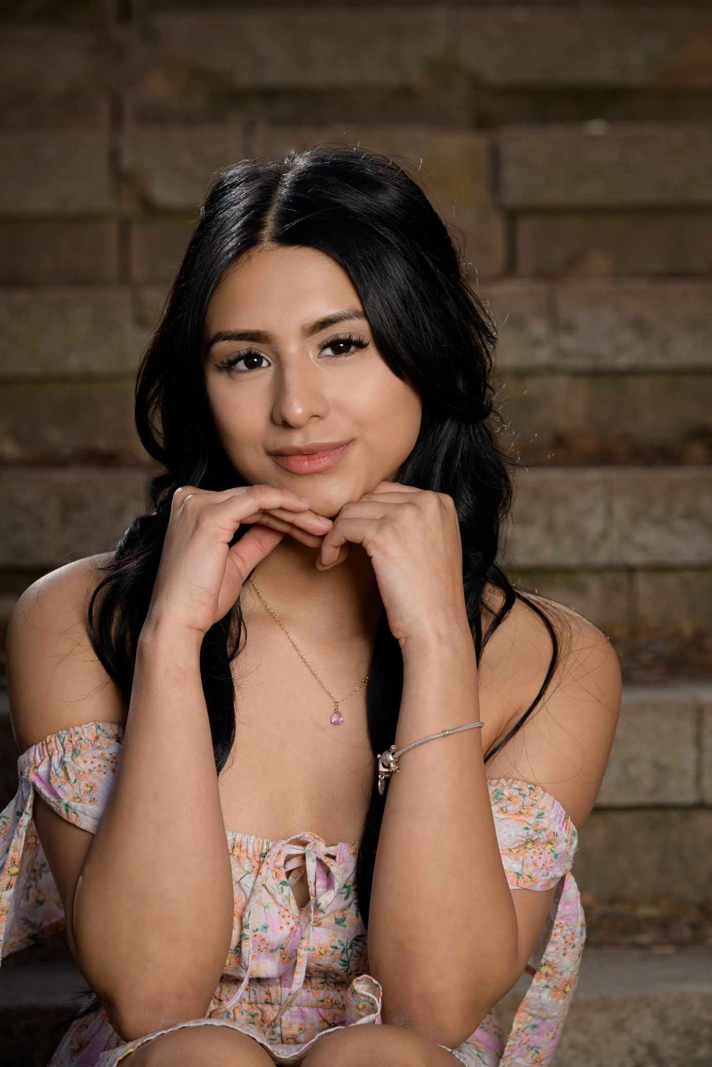 Beautiful young woman with dark hair in floral off-shoulder dress sitting on stone steps.
