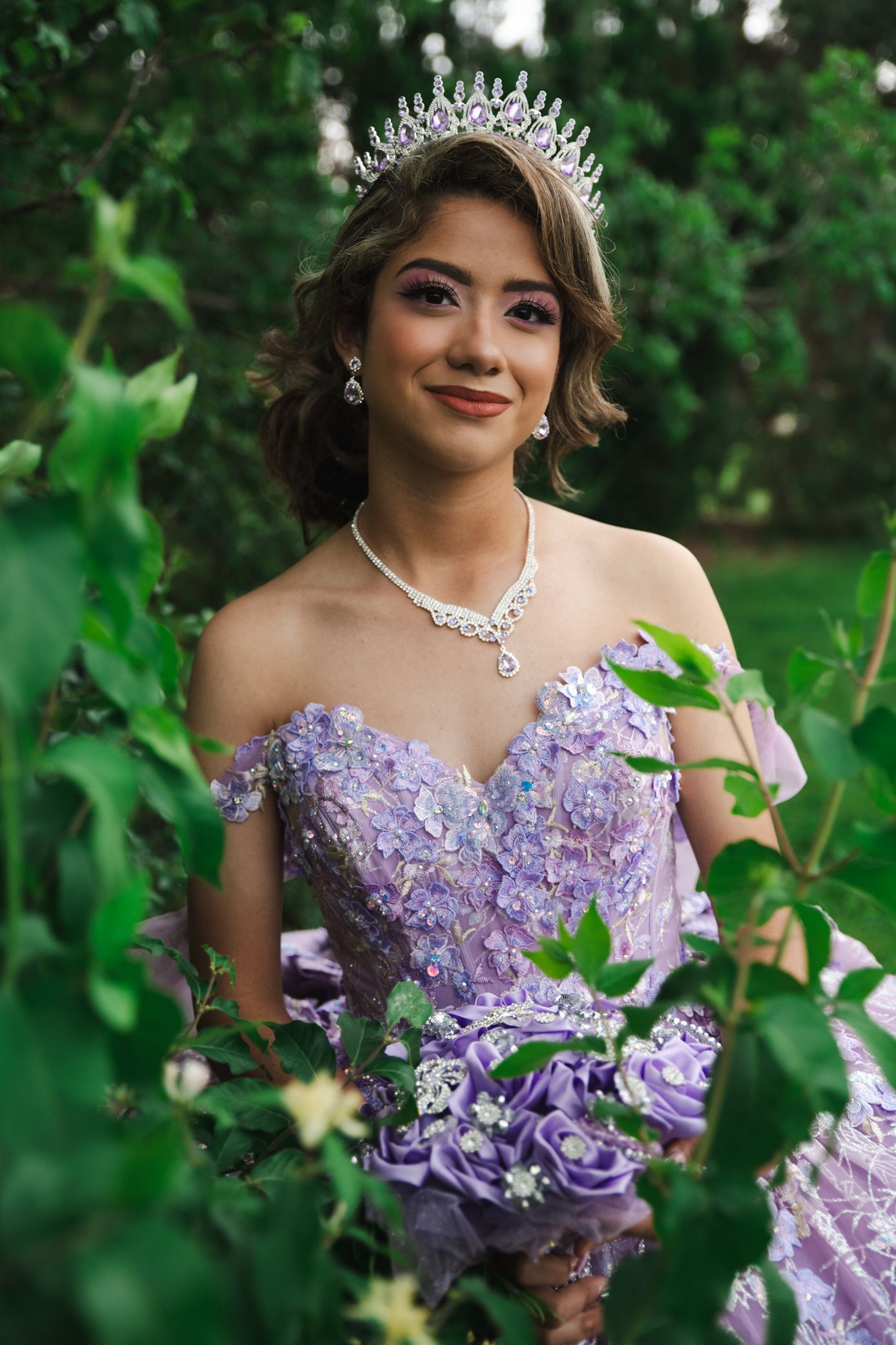 Young woman in lavender quinceañera gown with crystal tiara, pearl necklace, and purple floral bouquet outdoors.